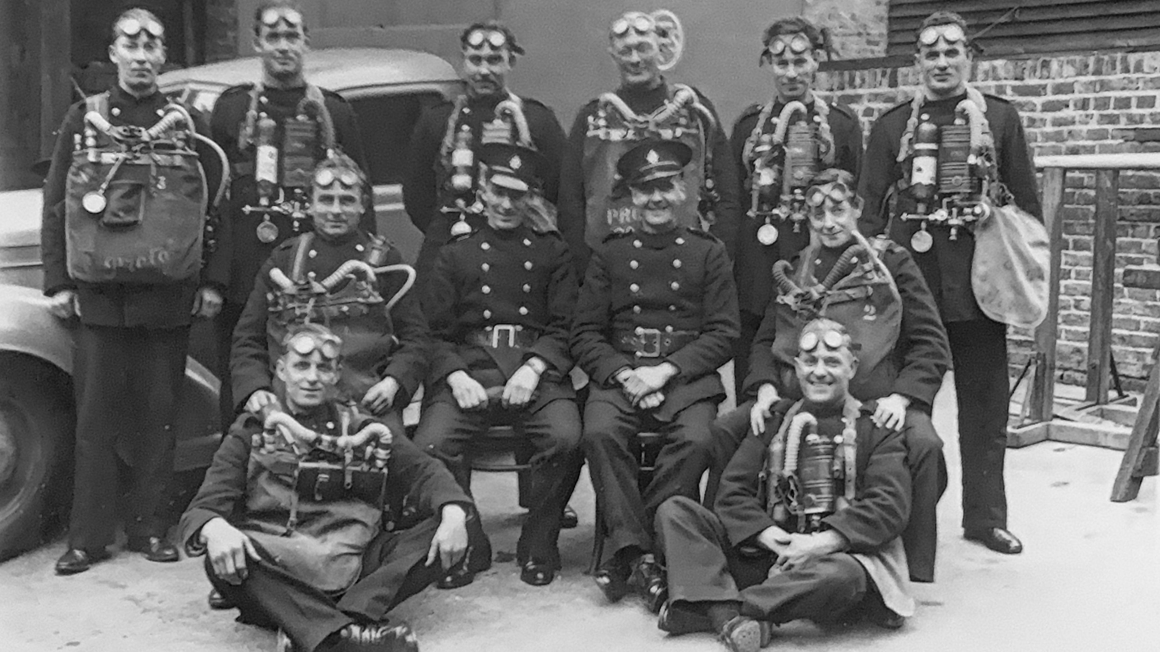 A group of firefighters posing for a photo with some standing at the back, some on chairs and two seated cross legged on the floor at the front. They are all looking at the camera and wearing a lot of safety gear as well as button up jackets.