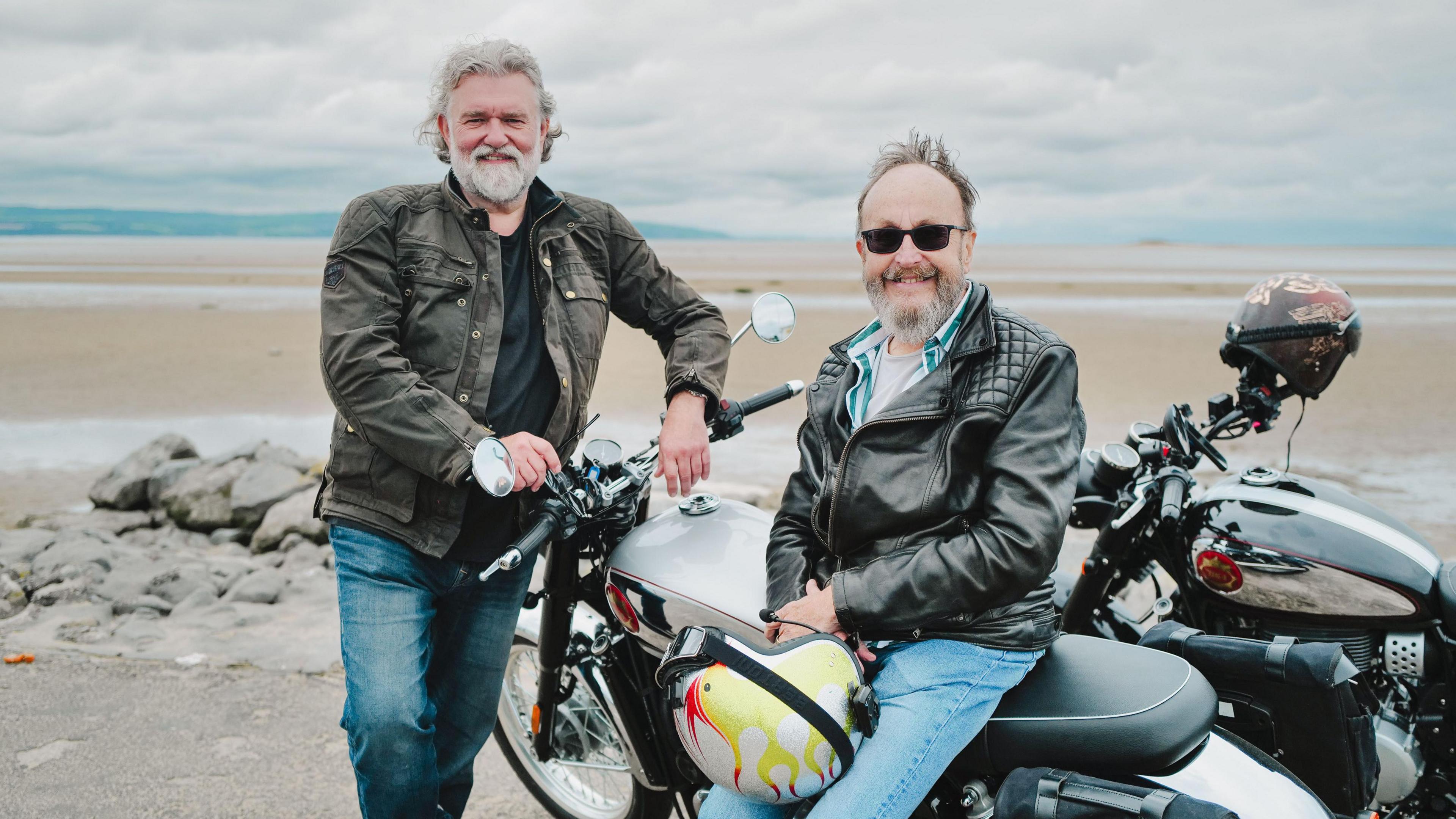 Si King is a man in his 50s wearing a dark jacket and jeans with a white beard and hair standing on a beach in front of a motorbike. Sitting on the bike is Dave Myers who is a similar age and wearing a leather jacket and jeans with a motorbike helmet in his hands