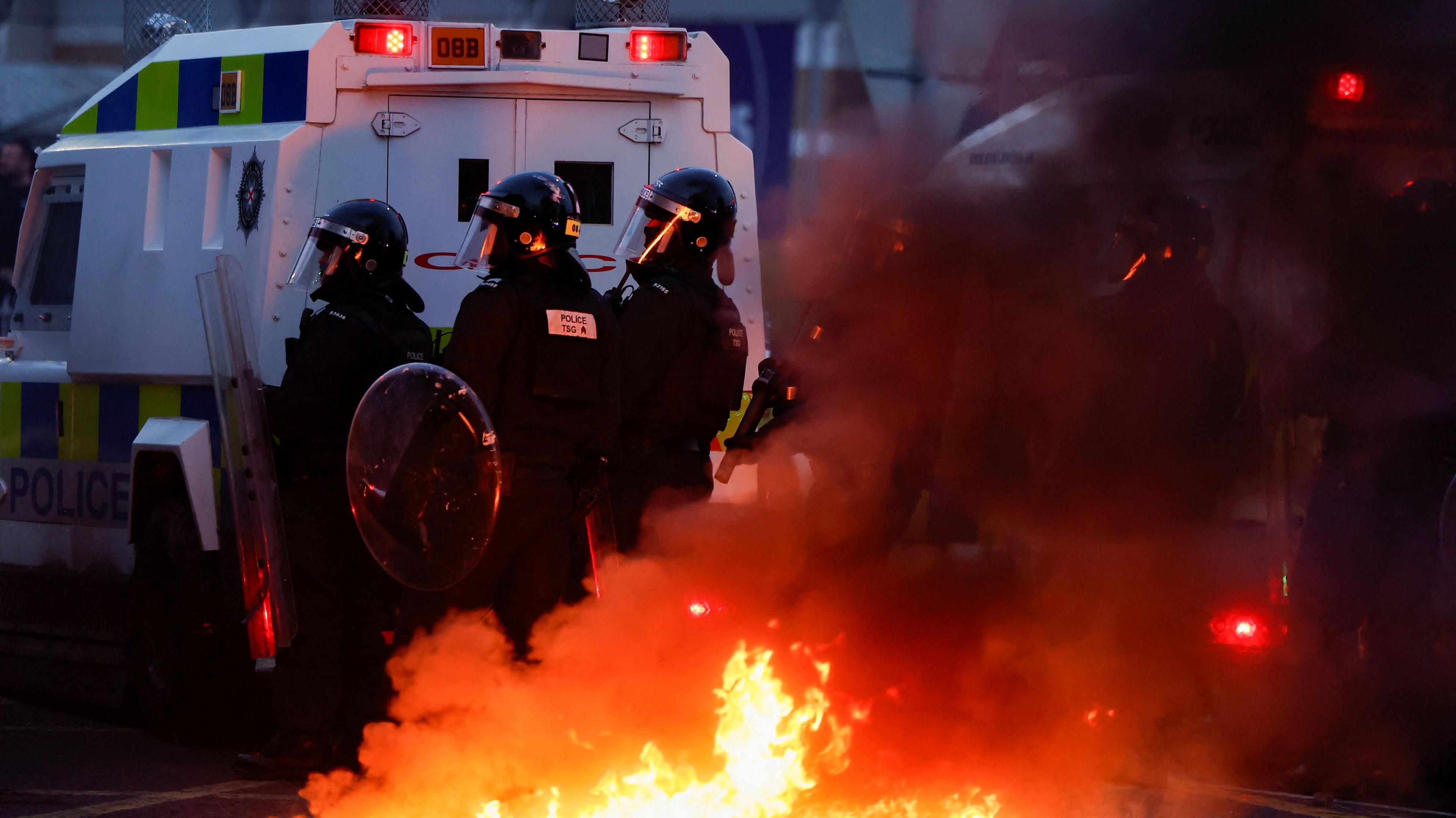 Armed police officers wearing a black uniform standing behind a white police jeep. There is flames rising into the air behind them.