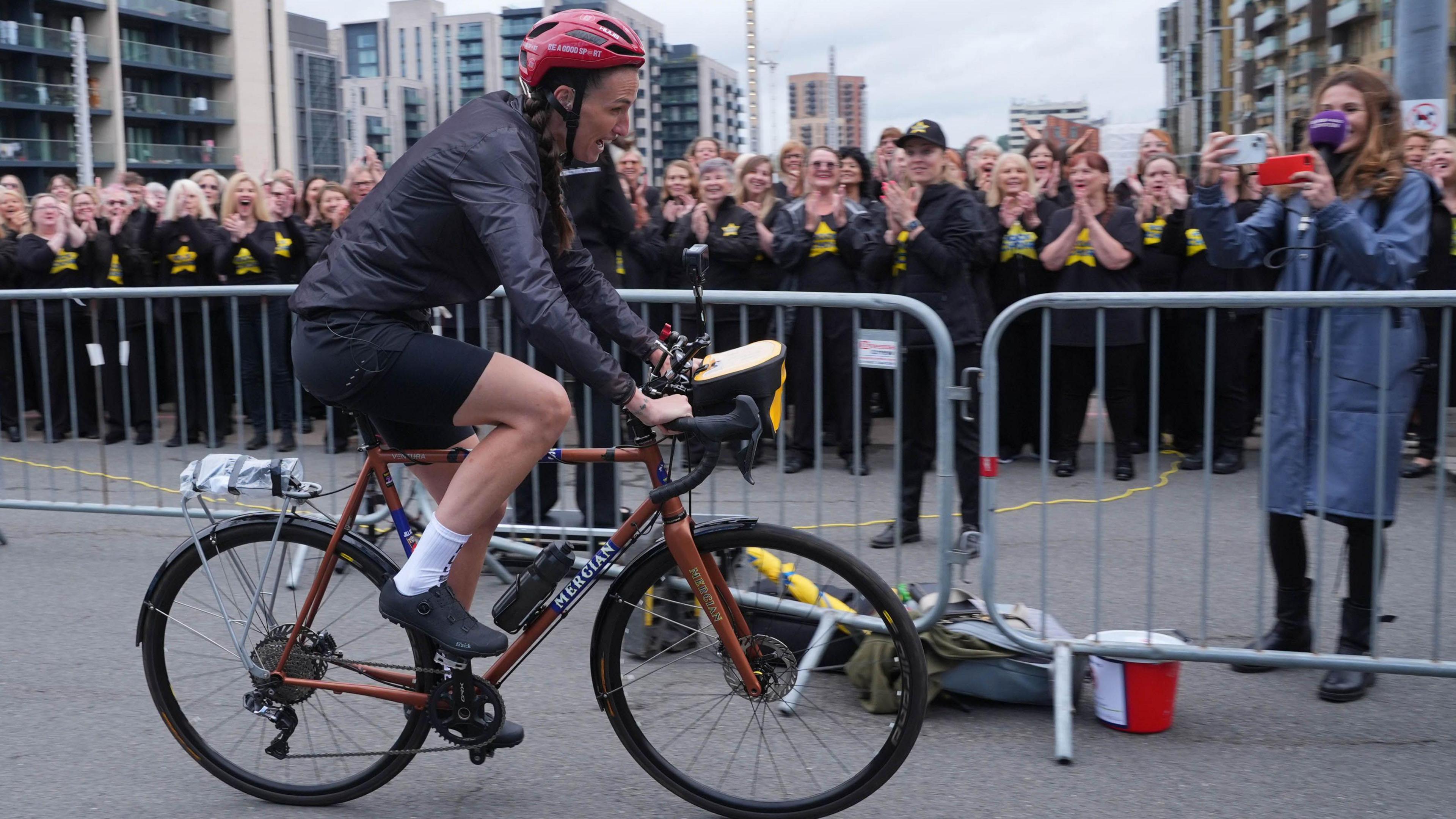 Jill Scott on her pushbike in red helmet and black cycling gear, a long sleeved waterproof top, her dark brown hair in a long plait as crowds in a line are cheering behind the barrier