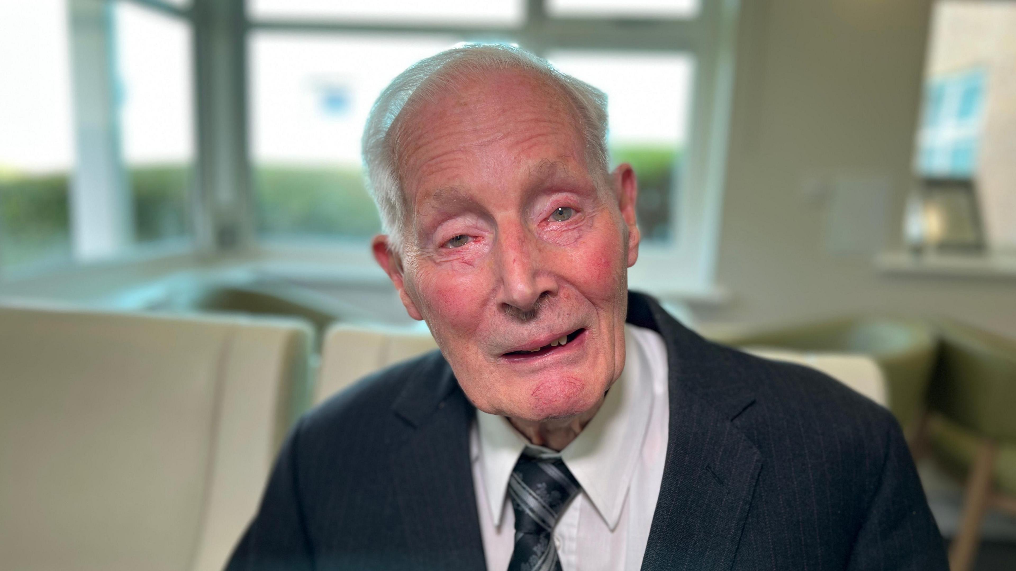 An elderly person in a dark suit, white shirt, and patterned tie sits in a room with large windows and visible furniture.