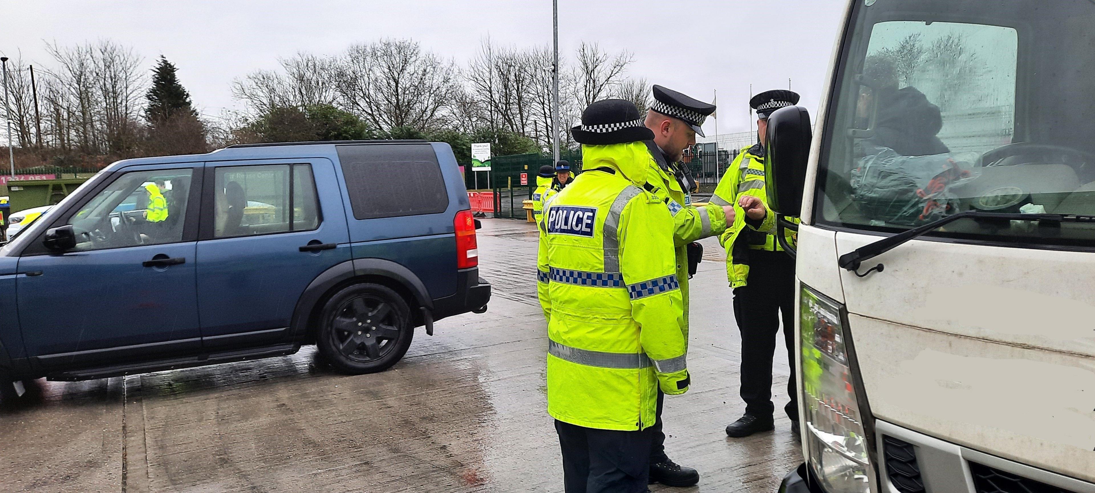 Three police officers, in high-viz jackets, by a large white van, talking to a person, in a car park, by a blue vehicle. The ground is wet and other officers can be seen in the distance. 