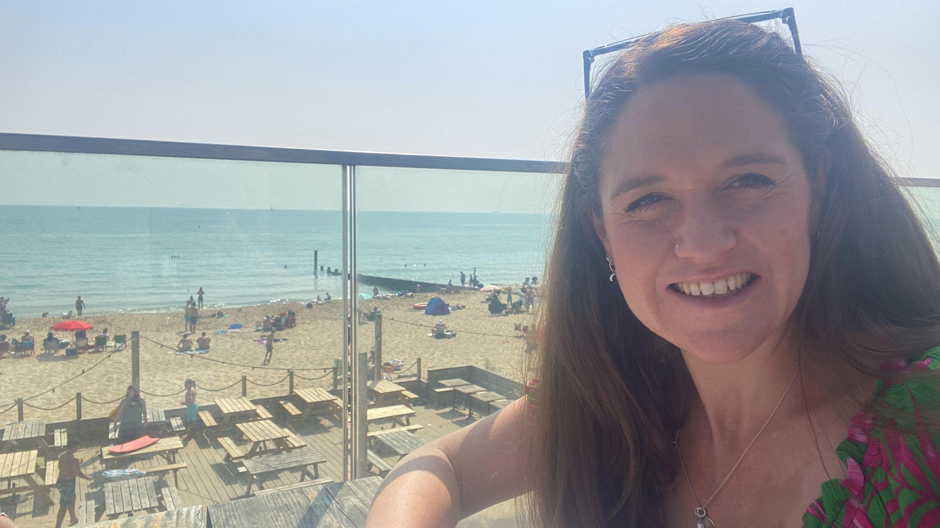 A woman in a selfie image on a balcony overlooking a beach on a sunny day. She has long dark hair with sunglasses resting on top of her head. She is wearing a green and pink top.