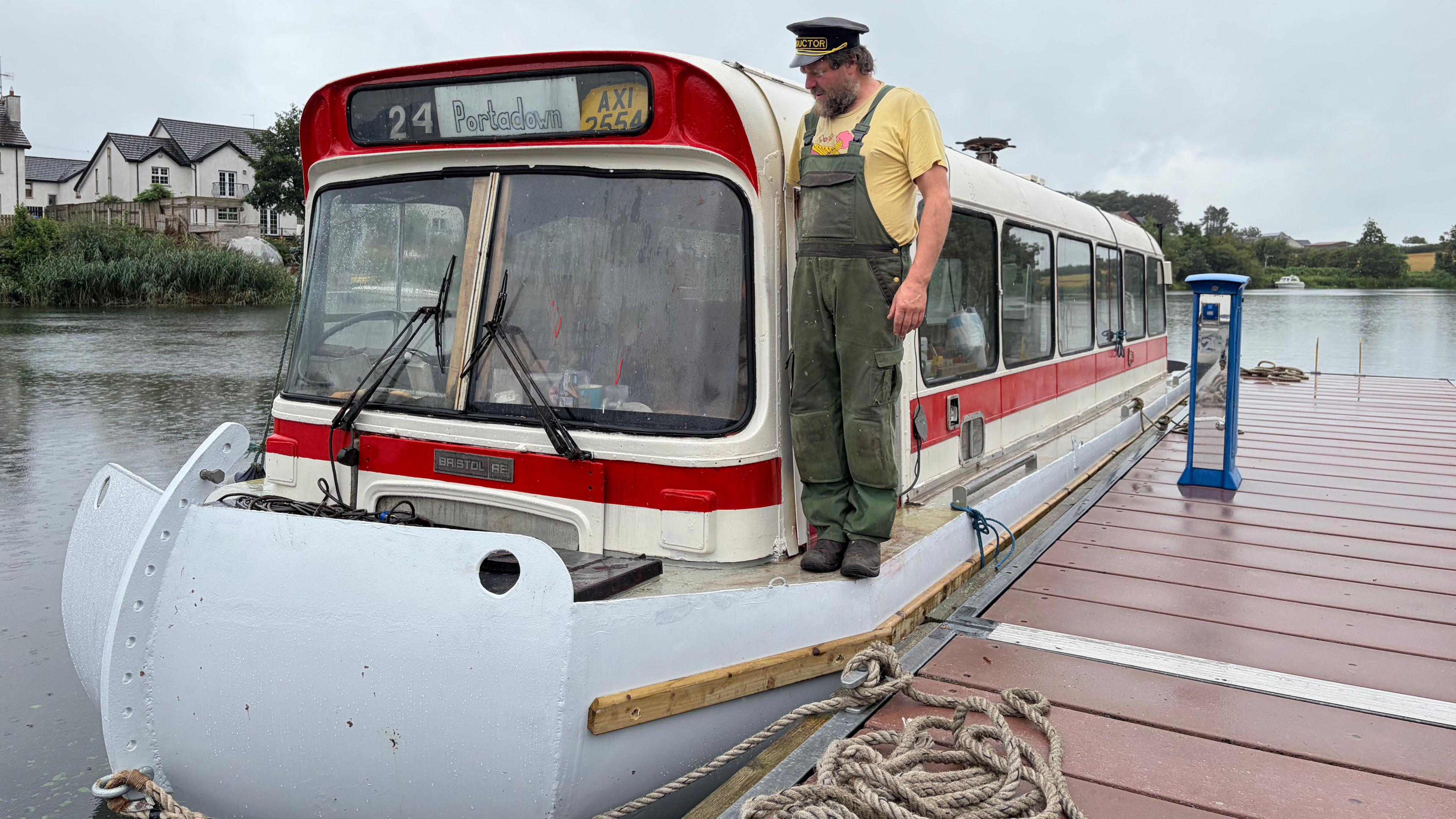 Paddy Bloomer stands on the deck of his bus boat. He is wearing green dungarees over a yellow T-shirt and a black peaked cap with "conductor" written on it in gold font. He has a bushy beard. The bus's destination sign reads "24 Portadown" and beside it is a licence plate reading "AXI 2554".