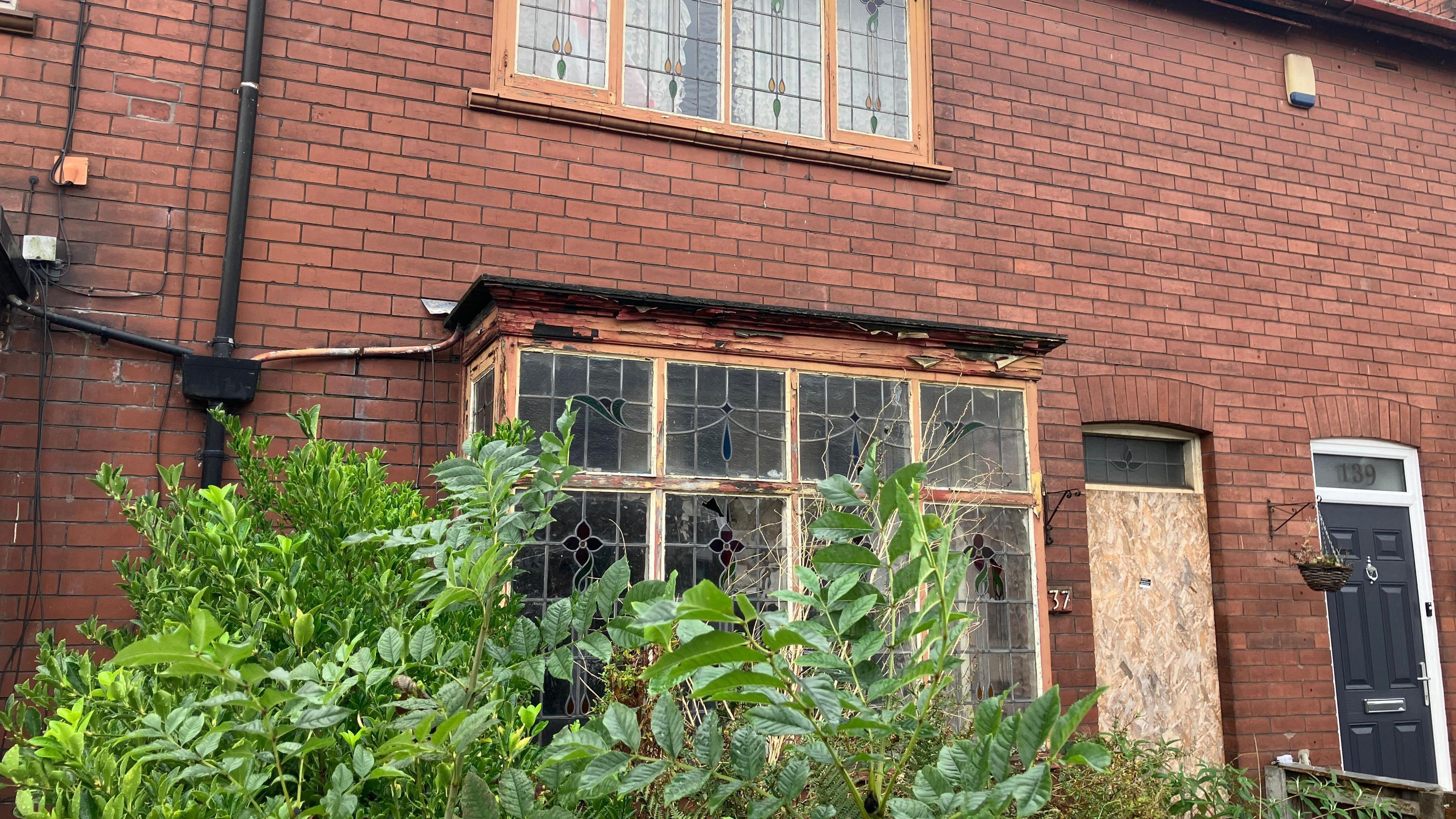 Empty home in Wigan with overgrown hedges and foliage, a boarded up door and old damaged windows