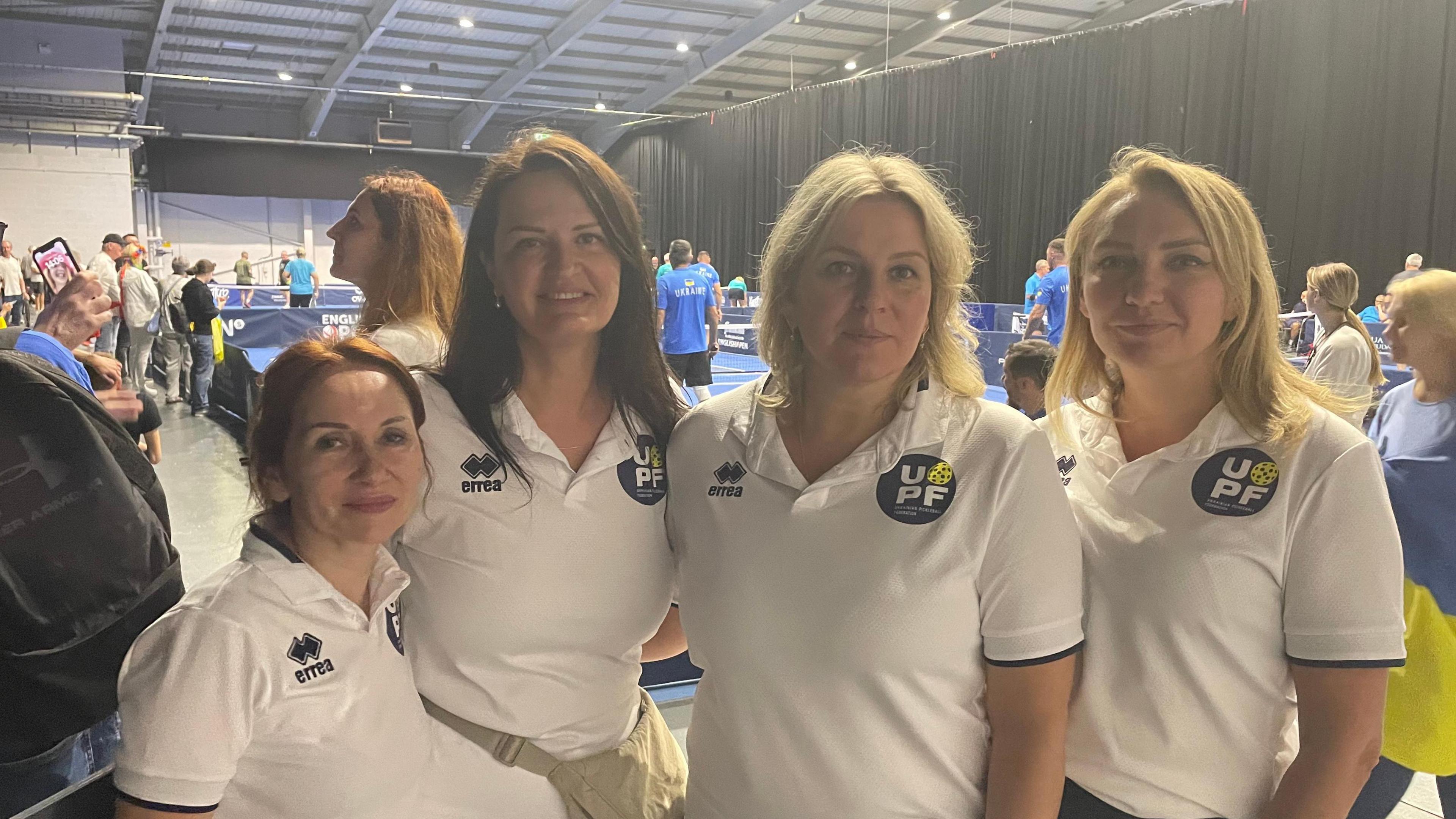 Three women wearing white T-shirts in front of a pickleball court