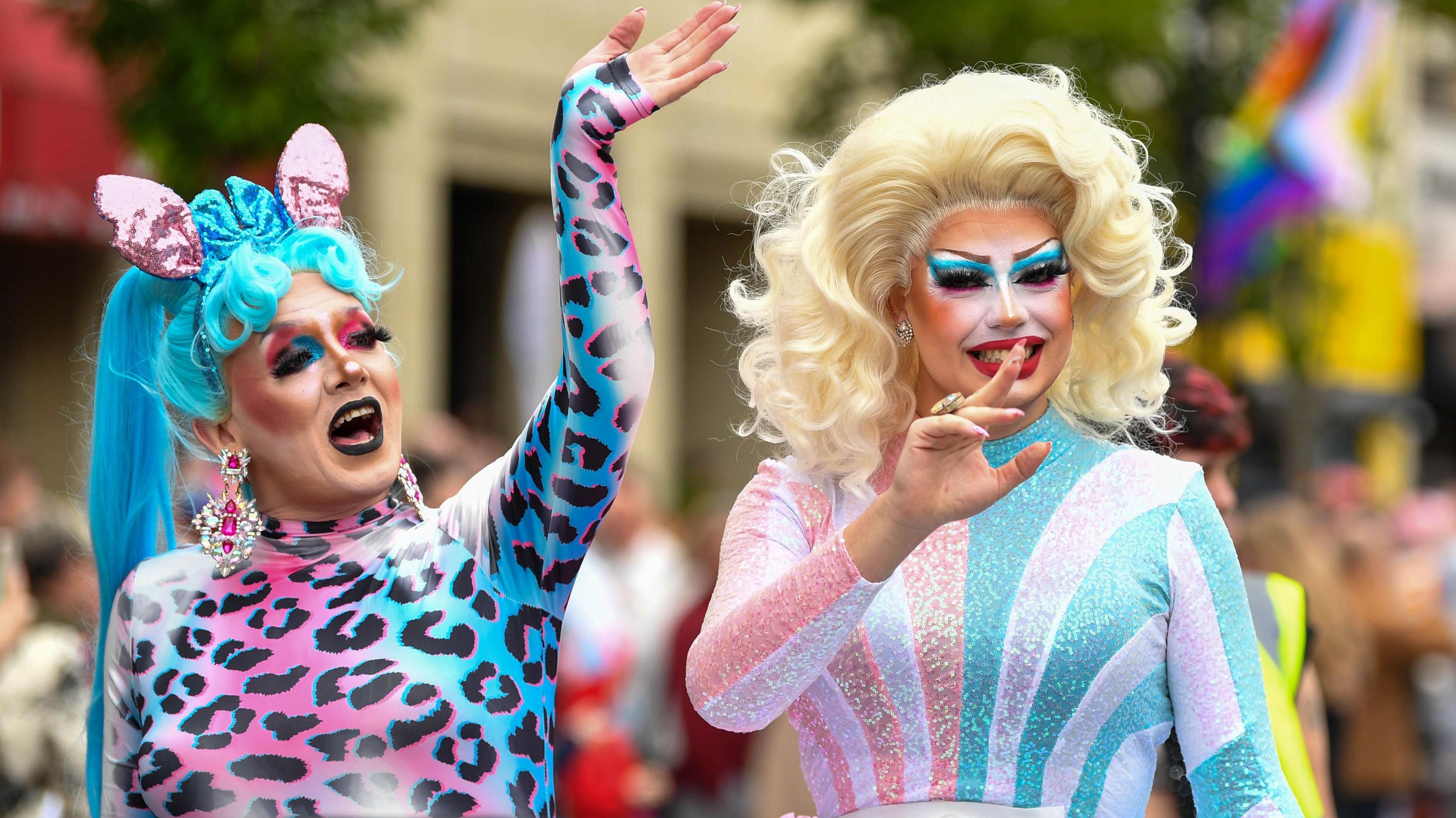 Two people dressed in bright colours on a float at a festival. One is wearing a blue wig and wearing pink and blue cheetah print and big silver and pink earrings. The other is wearing a blond wig and wearing pink, blue and silver. Both are wearing make-up.