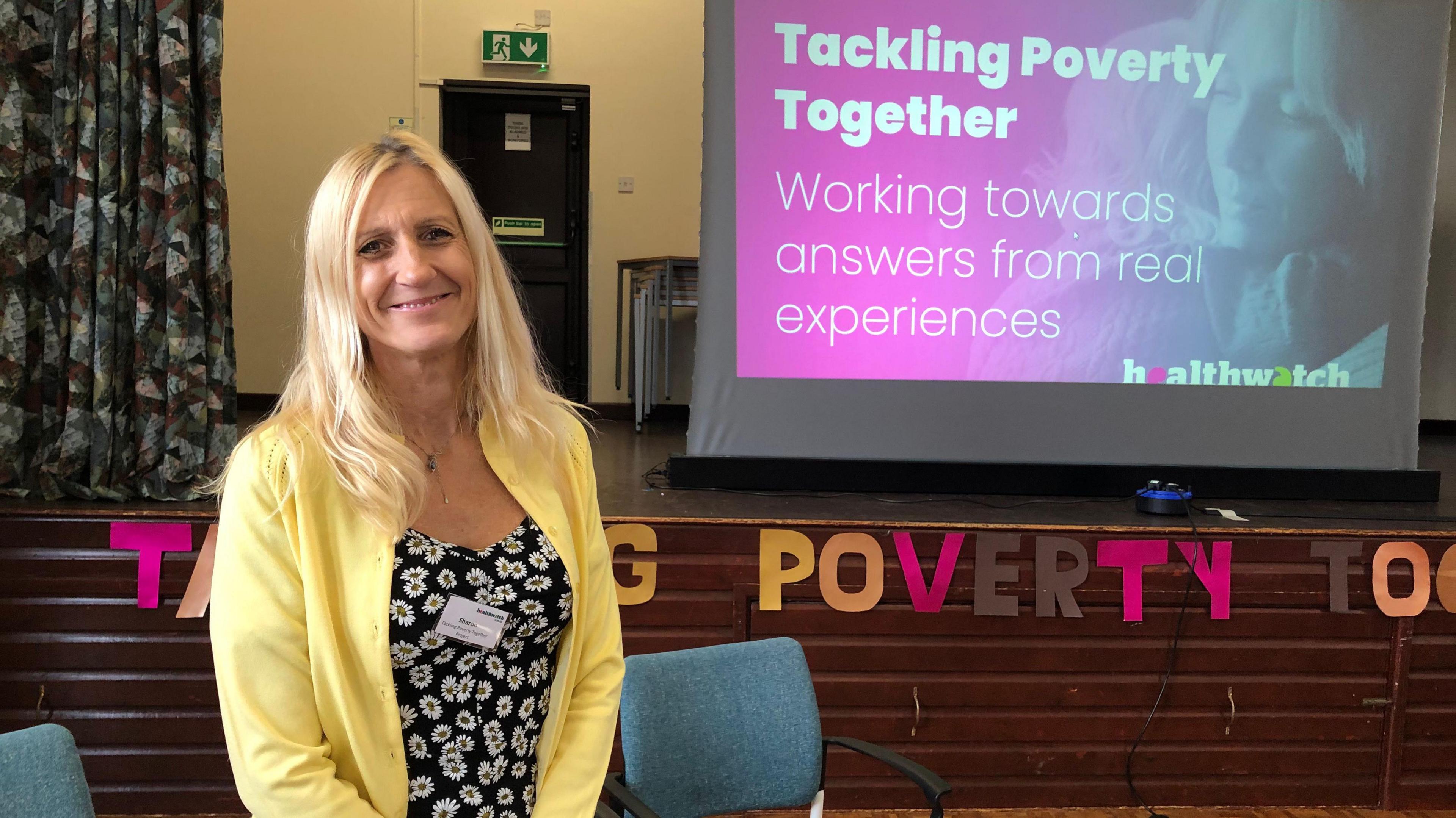 Sharon Cony stands in front of a projector screen in a village hall which has a title of Tackling Poverty Together