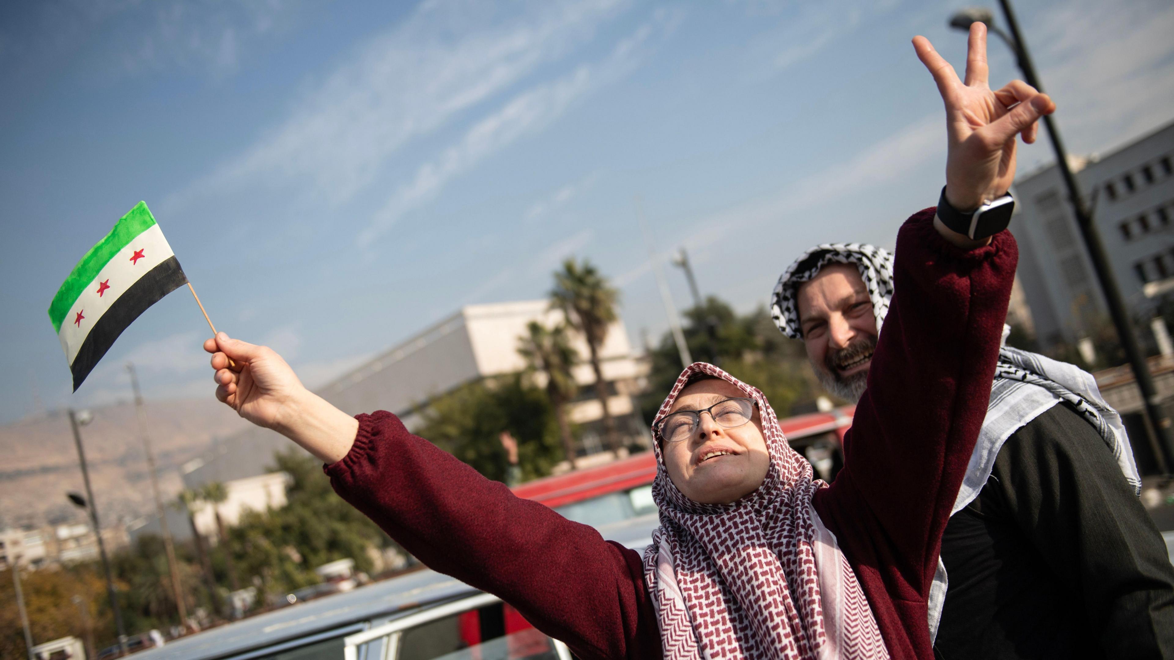 A man and a woman celebrate in the street after Syrian rebels captured the city of Damascus. The woman raises her hands to the sky - one of her hands is holding a Syrian flag.