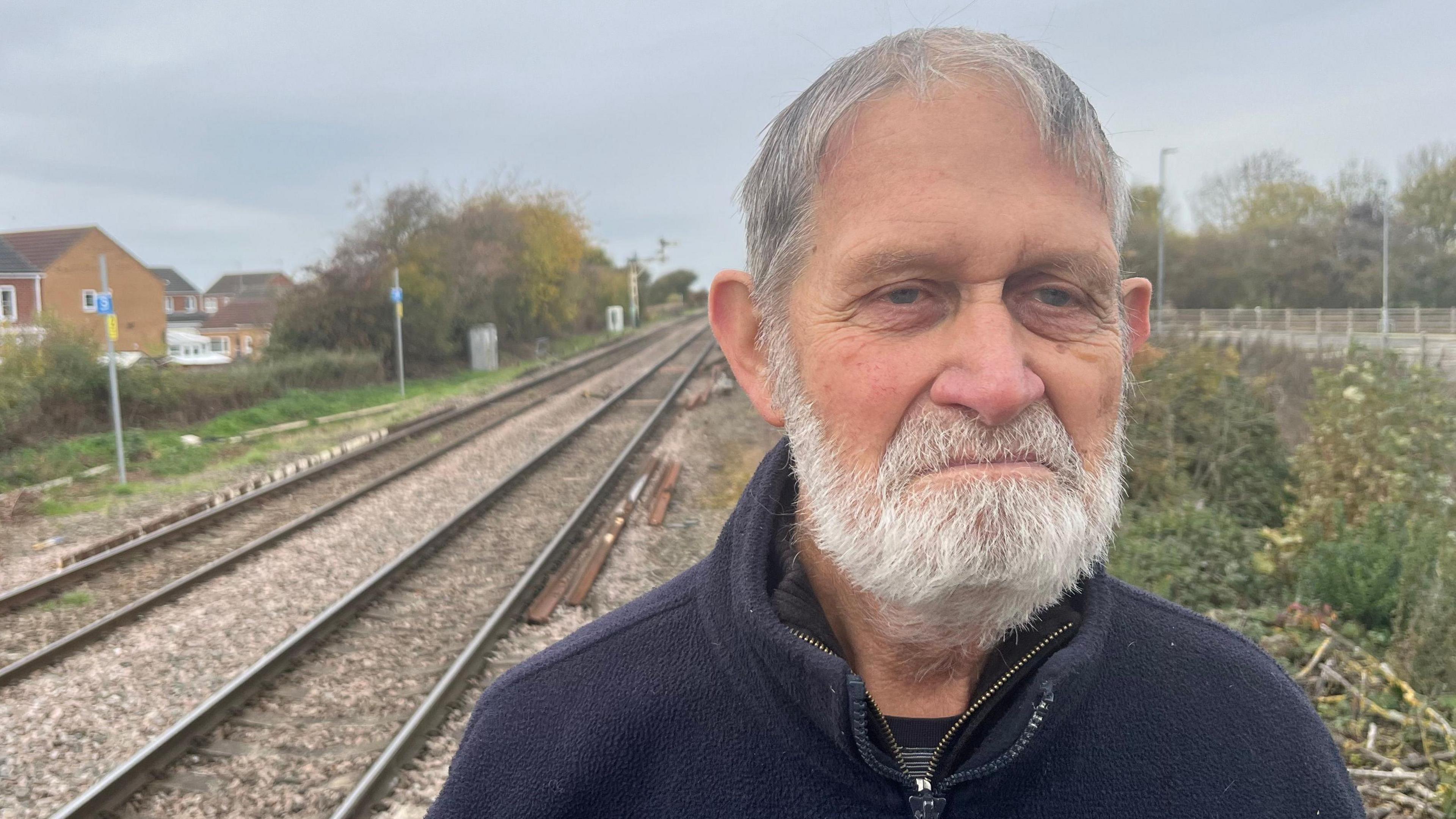 Peter Wright, 77, has short grey hair with a matching beard and moustache, he is wearing a blue fleece top with a striped t-shirt underneath and is standing on a railway platform with the tracks behind him, a semaphore sign, which is black and white and is suspended from a metal tower beside the track.