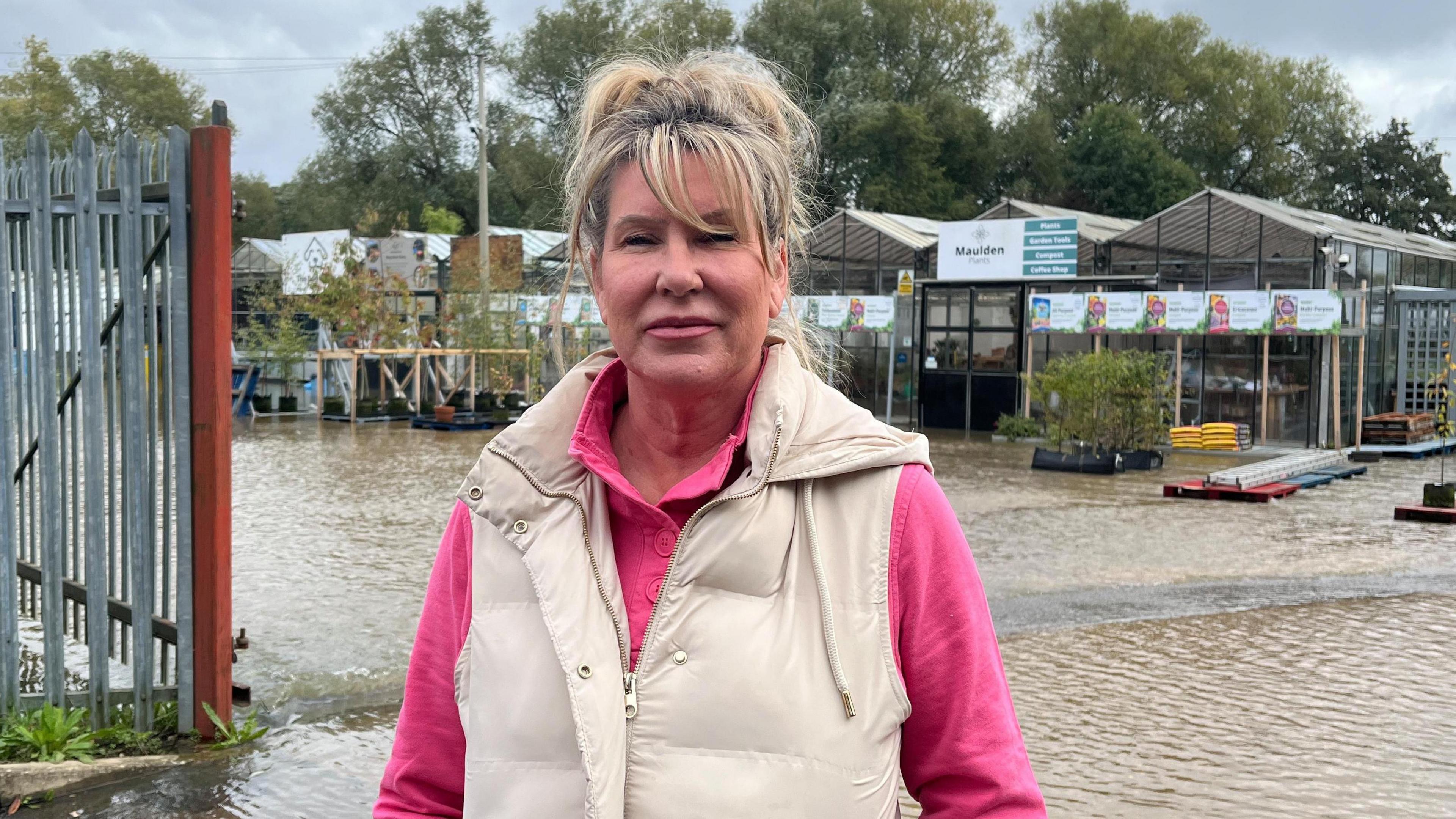 A woman who runs a doggy day care business on the site of the Maulden Garden Centre standing by the gate entrance with the flood area behind her. She is wearing a pink jumper under a cream sleeveless jacket.