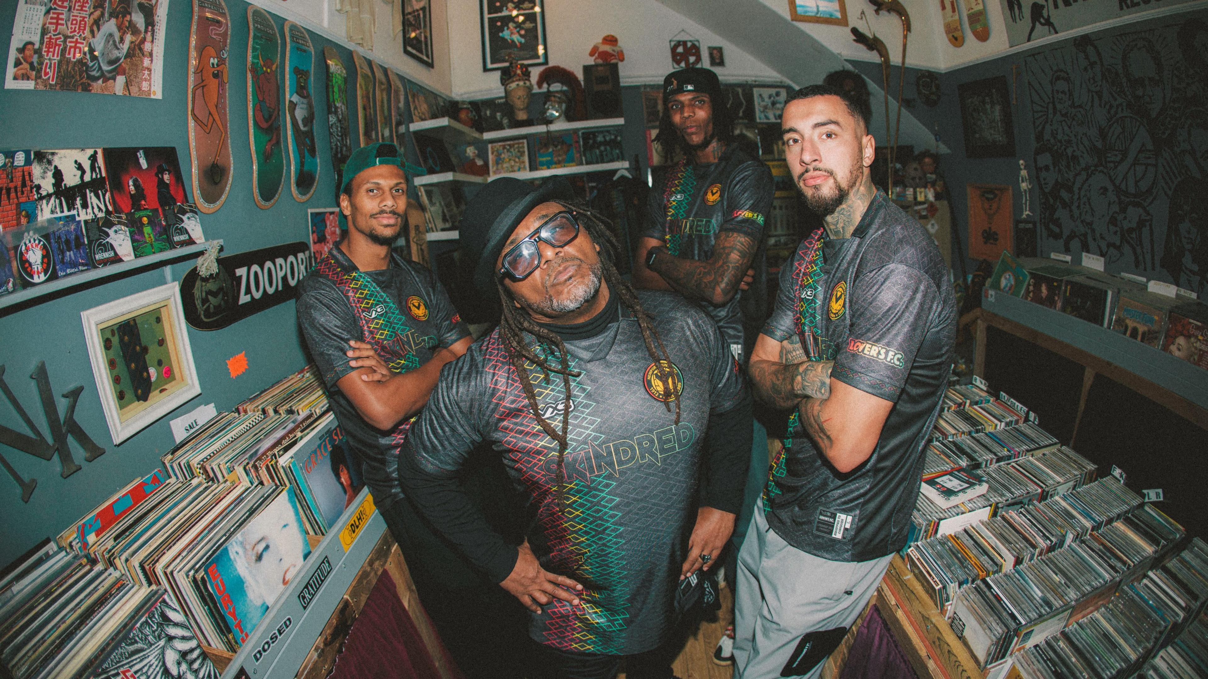 At front and centre, Skindred's lead singer Benji Webbe wears the new on-off kit design. He wears a black hat, black tinted glasses and has his hands on his hips. Other band members stand behind him within the record store, all with their arms crossed staring straight faced at the camera. 