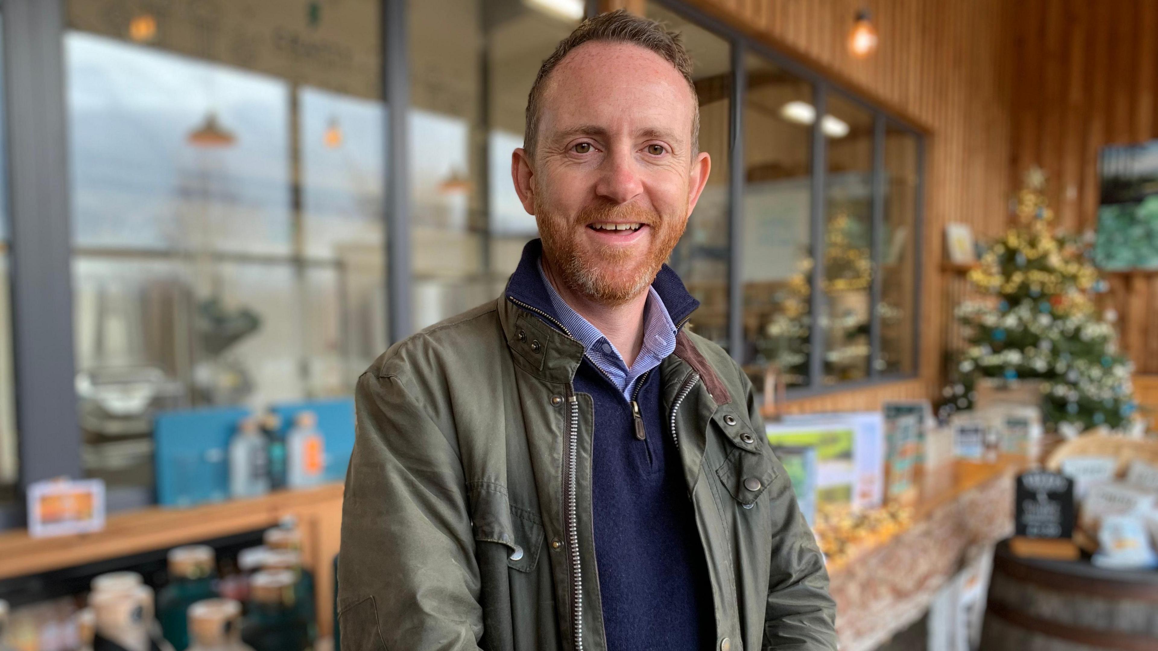 A red haired man with a beard in a green jacket stands in front of a wooden bench with a Christmas tree in the background