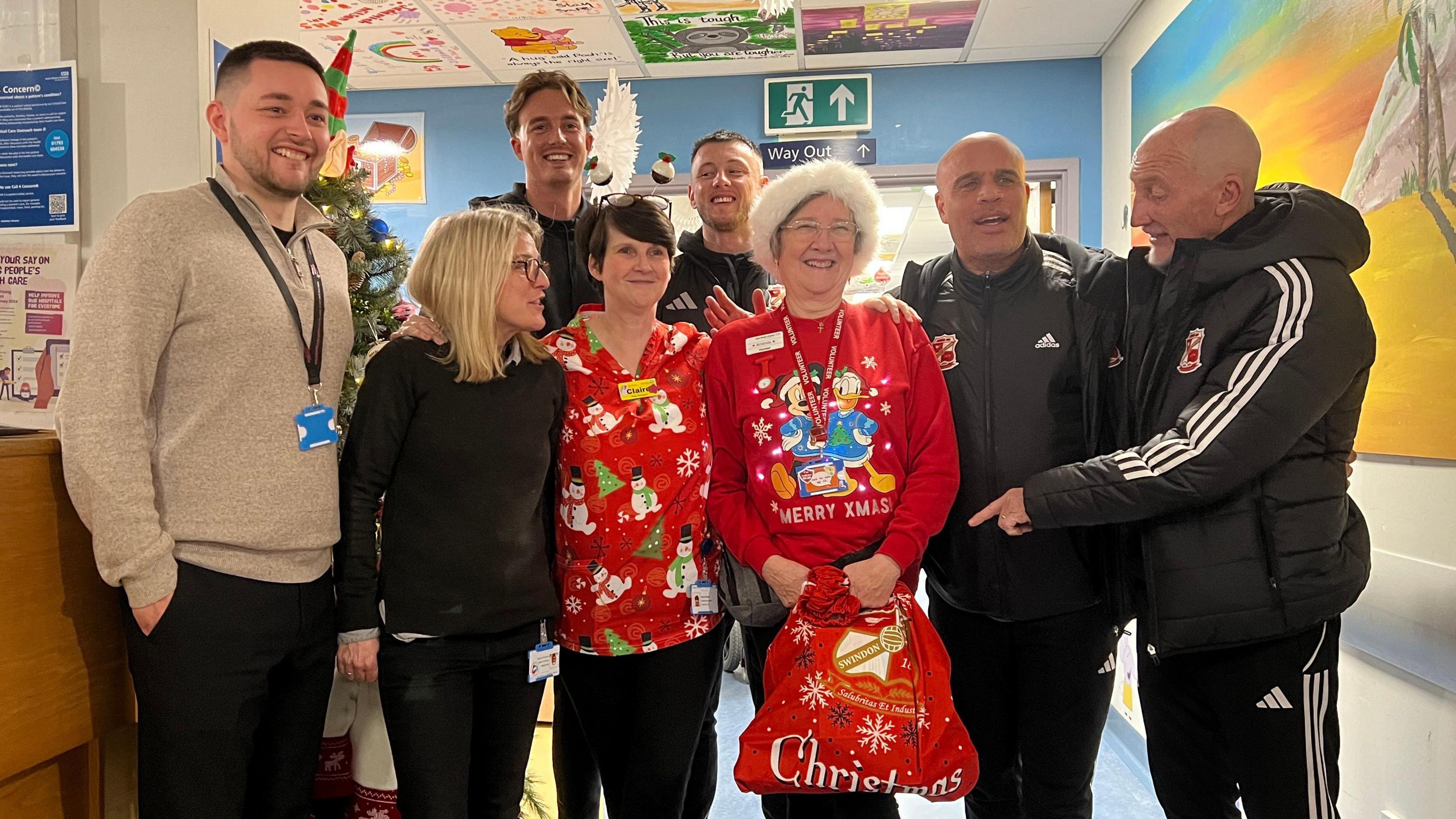 8 people standing for a photo in a hospital corridor - the women in the middle wearing christmas jumpers and a bald man pointing on the right to the Christmas bag held by one of the women