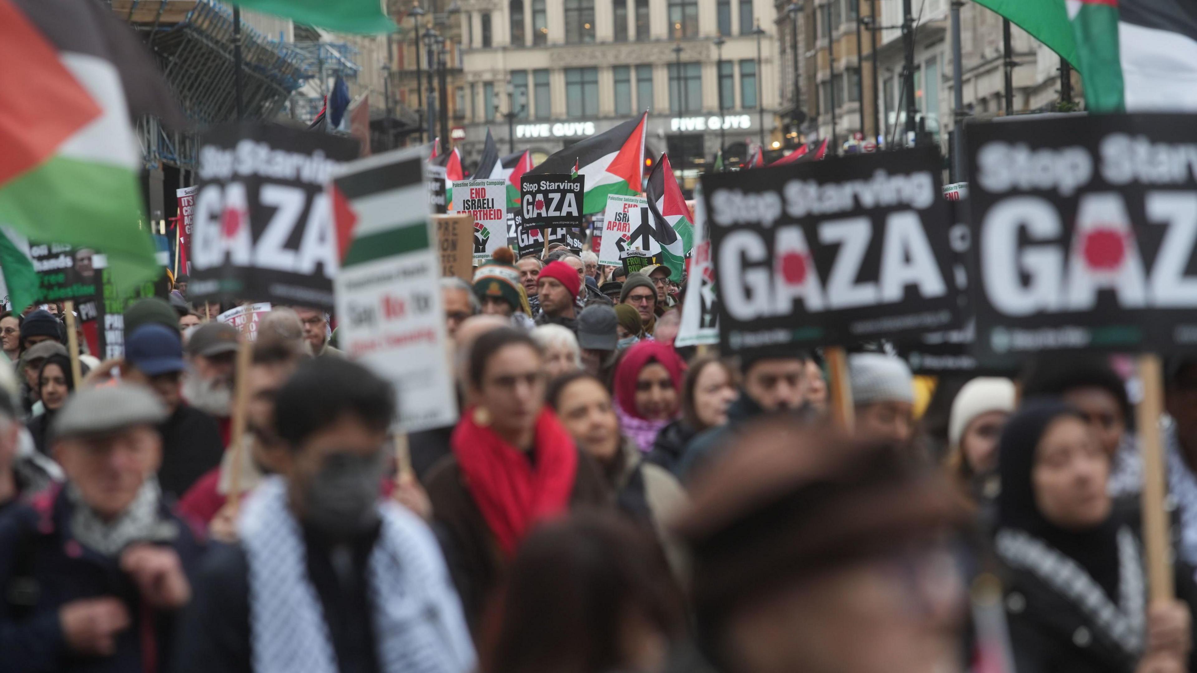 A large crowd of protesters marching through a street in London. Many hold placards that read "Stop Starving Gaza" and Palestinian flags are being waved above the crowd.