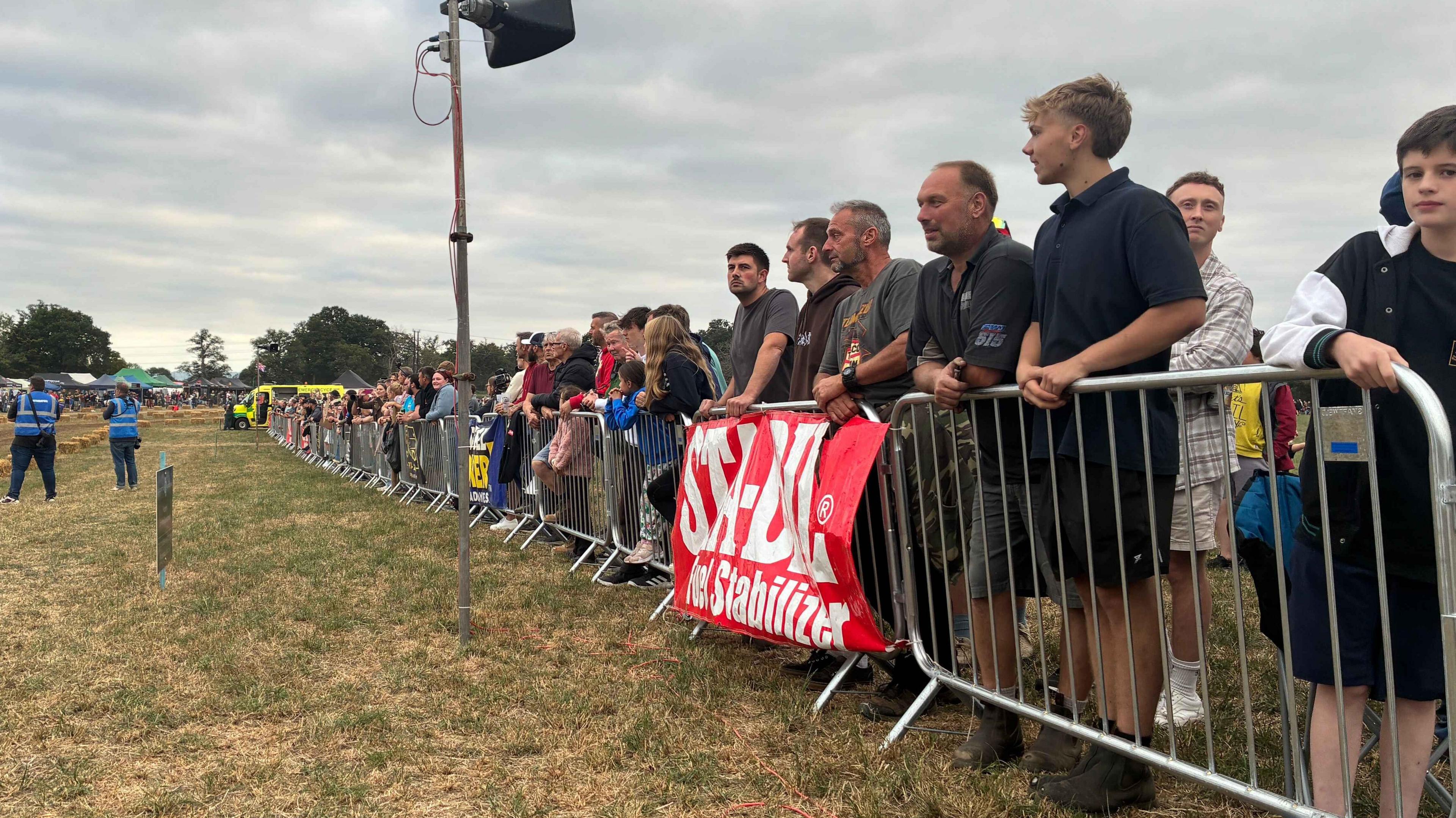 A large crowd is stood along the metal barrier at the side of a dirt race track 