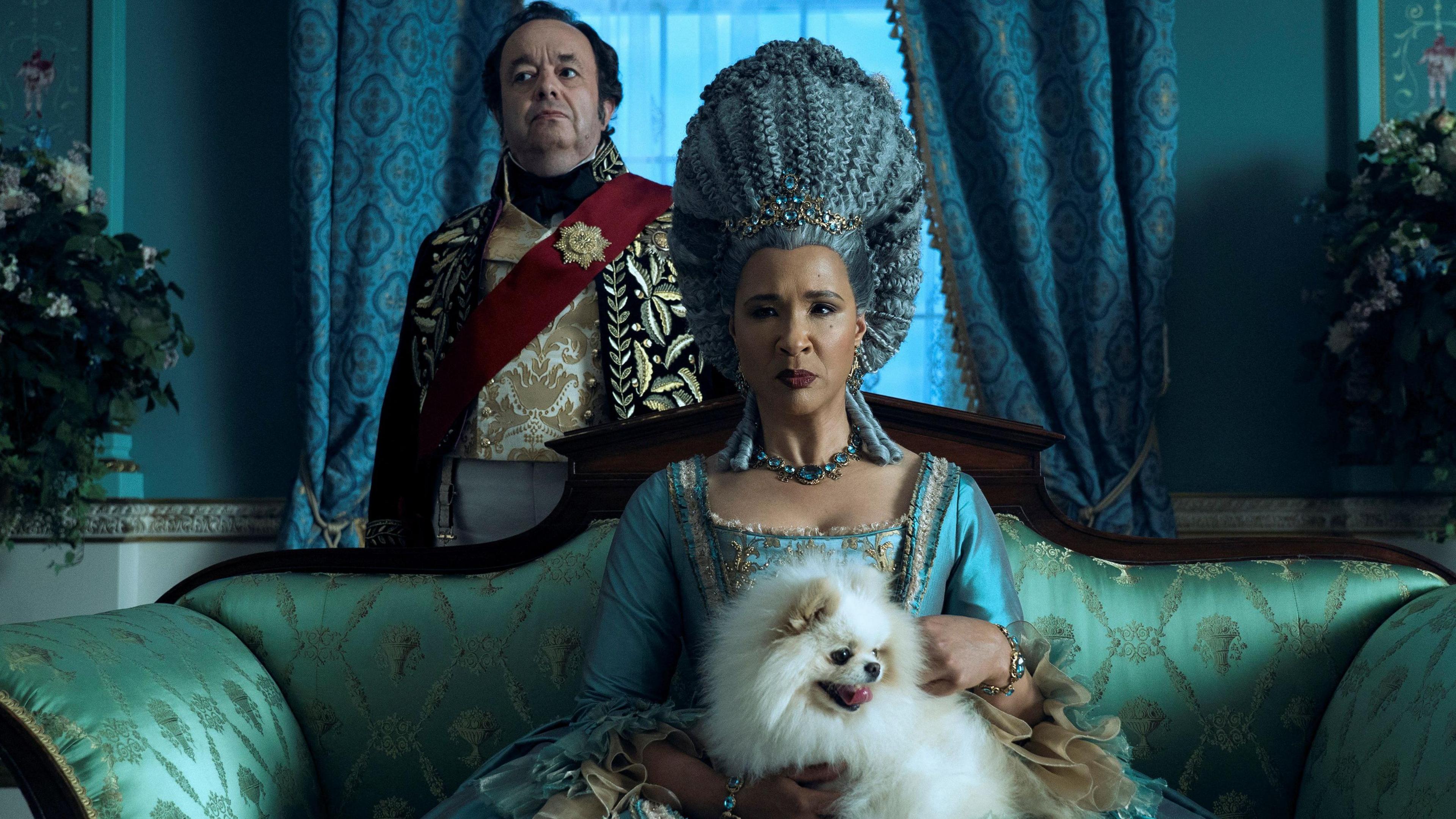 An image from the TV series Bridgeton with Golda Rosheuvel as Queen Charlotte sitting on an opulent green settee with a small white dog on her lap. She is wearing an intricate turquoise silk dress and her blue coloured hair is piled high on her head. 