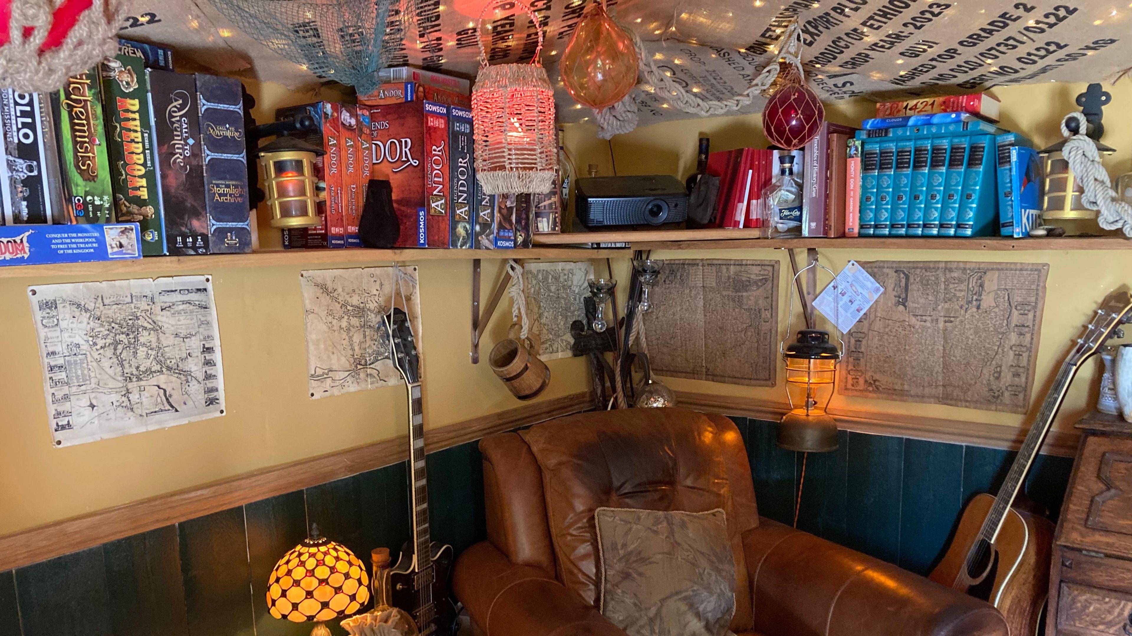 Another part of inside the shed. A brown leather chair rests in the corner with a guitar next to it. Above it a shelf that lines the wall and includes games and books on it.