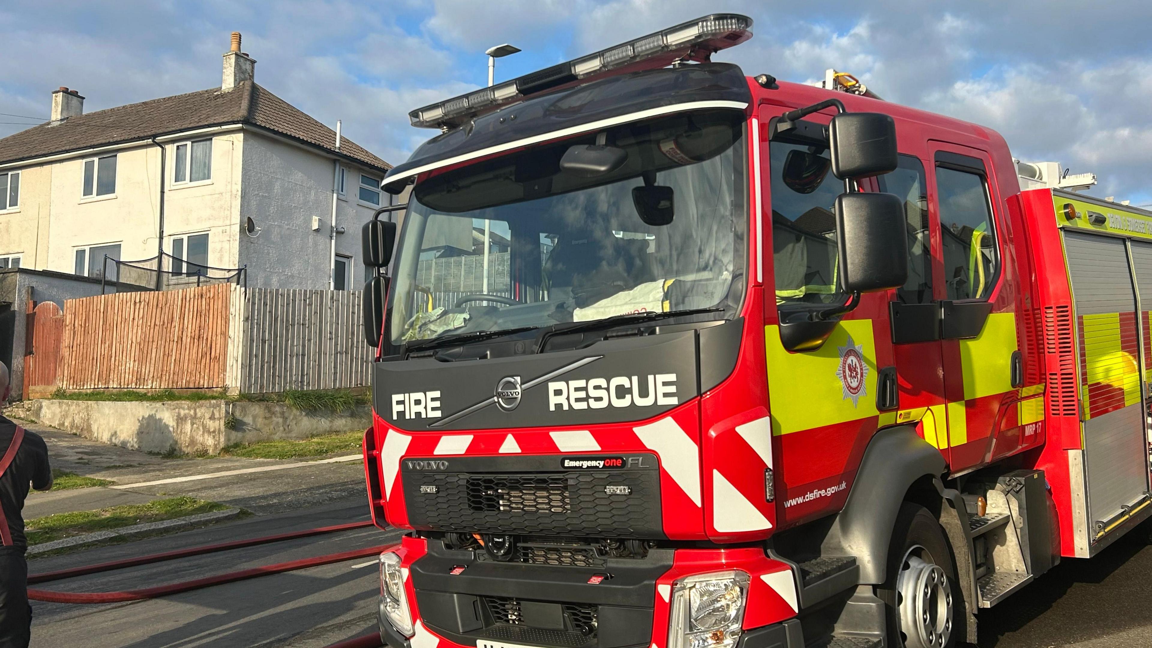 A red and yellow fire engine parked on a pavement. The front says 'fire rescue'. A house with a wooden fence in front of it is in the background.