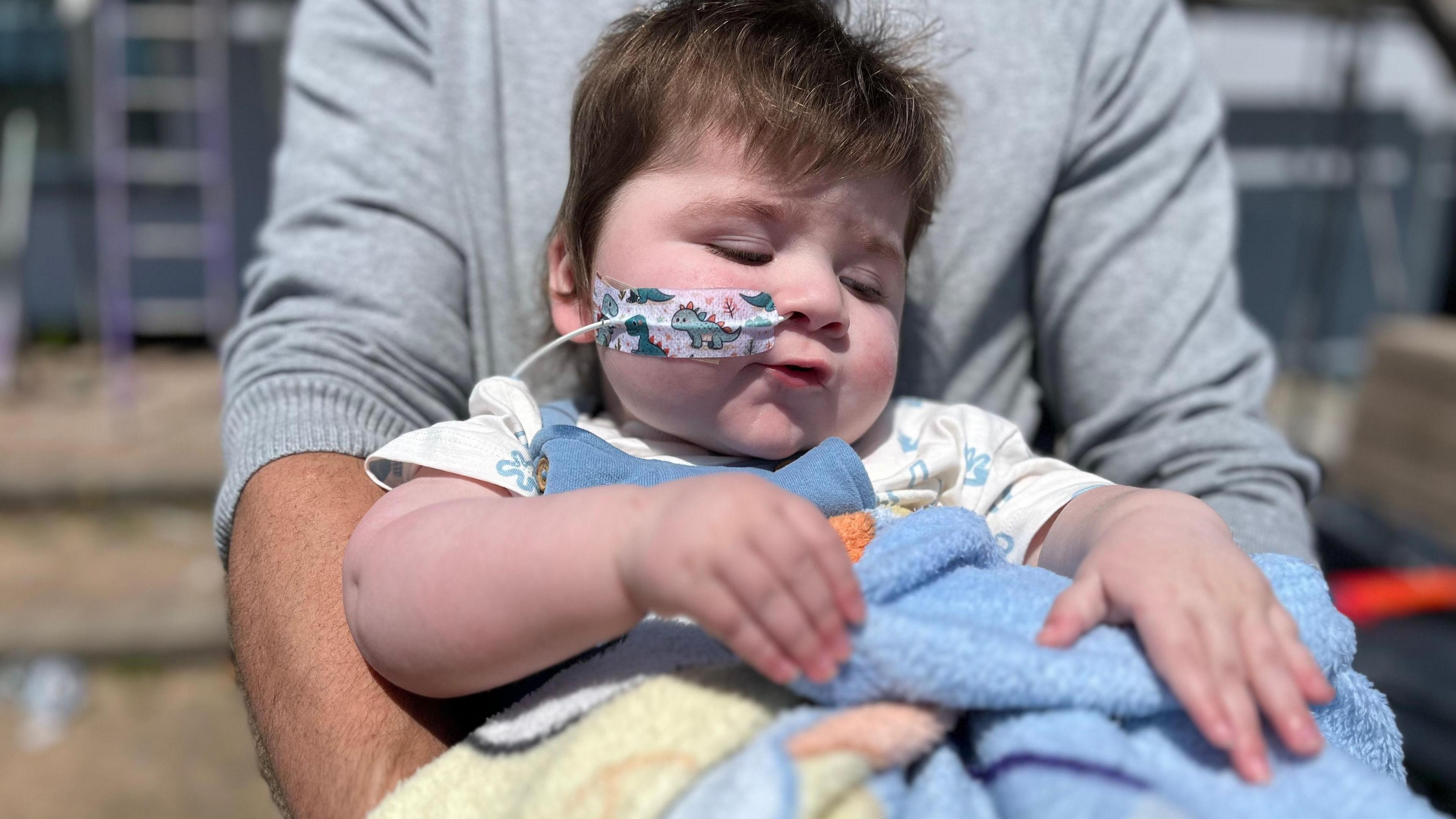 Jack being held by his dad Nick. Jack is in a blue baby grow and has a feeding tube going up his nose held in place with a dinosaur sticker. 