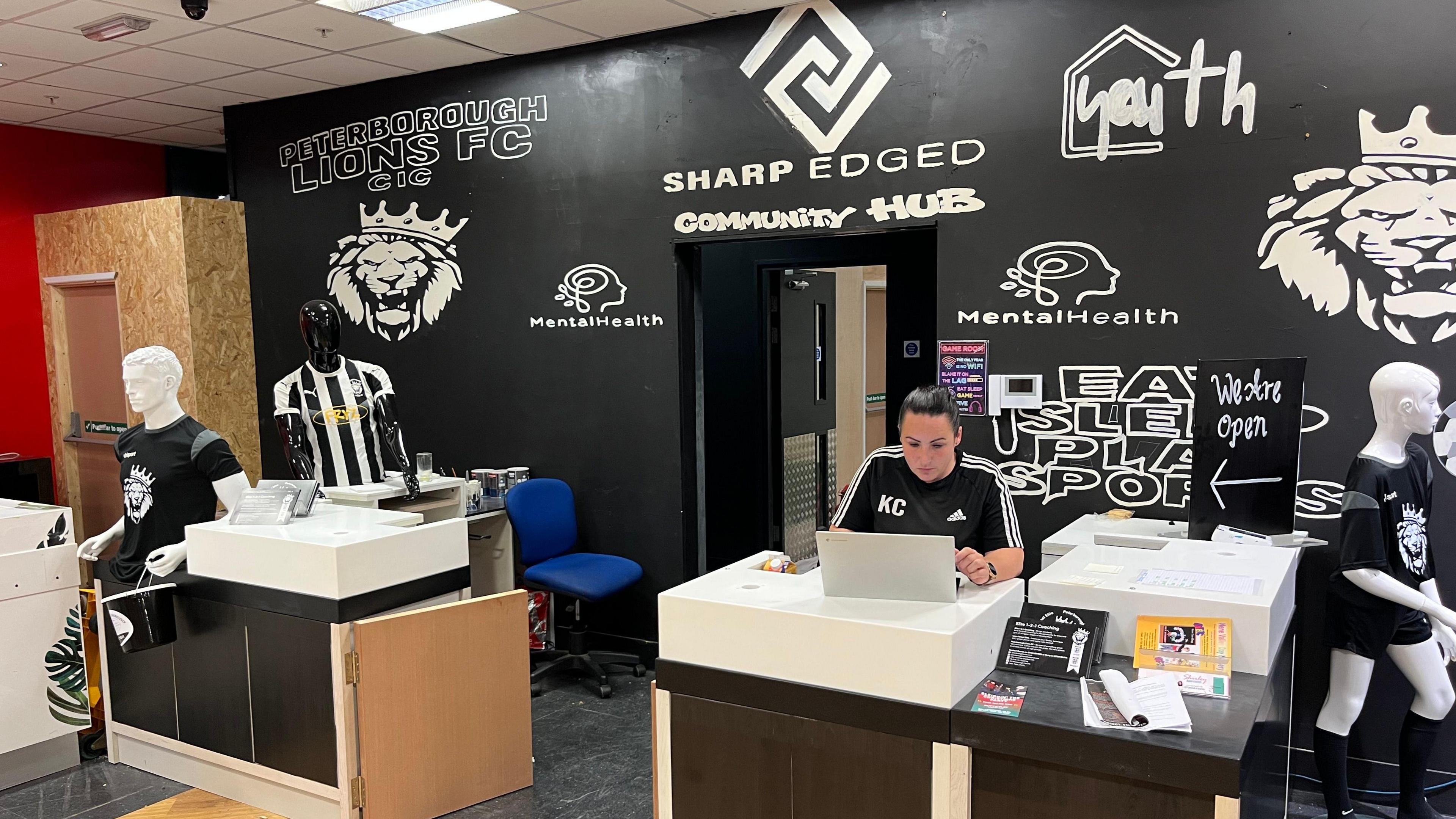 Kerry Cassidy is sitting at a desk and looking at a laptop. There is another desk and a space between the two leading into a doorway with 'Sharp Edged Community Hub' written above on the wall. There are other logos including a Peterborough Lions emblem (a drawing of a lion's face). There are mannequins wearing sports kits around the desks.