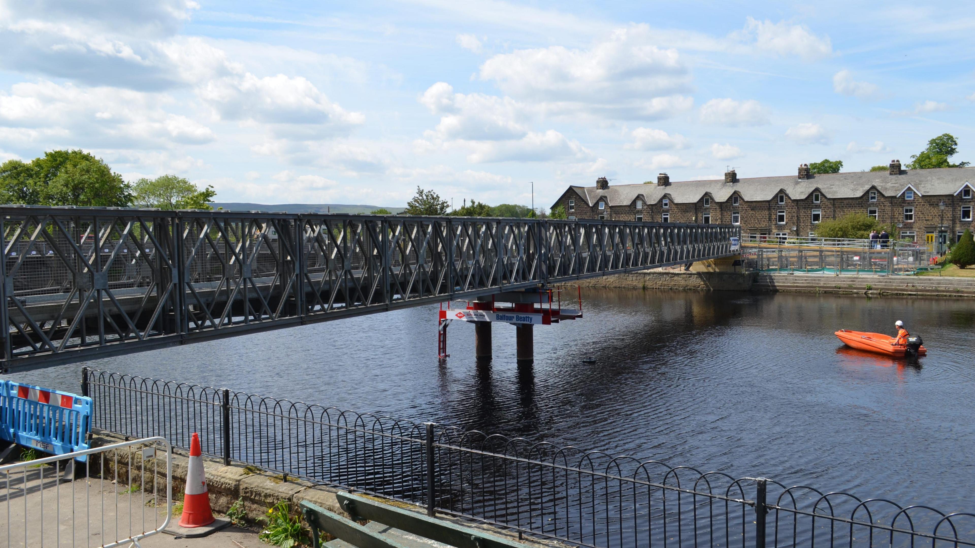 A footbridge running from one side of a river to another. A man in an orange kayak is on the water approaching it. Some houses can be seen on the other side of the river.