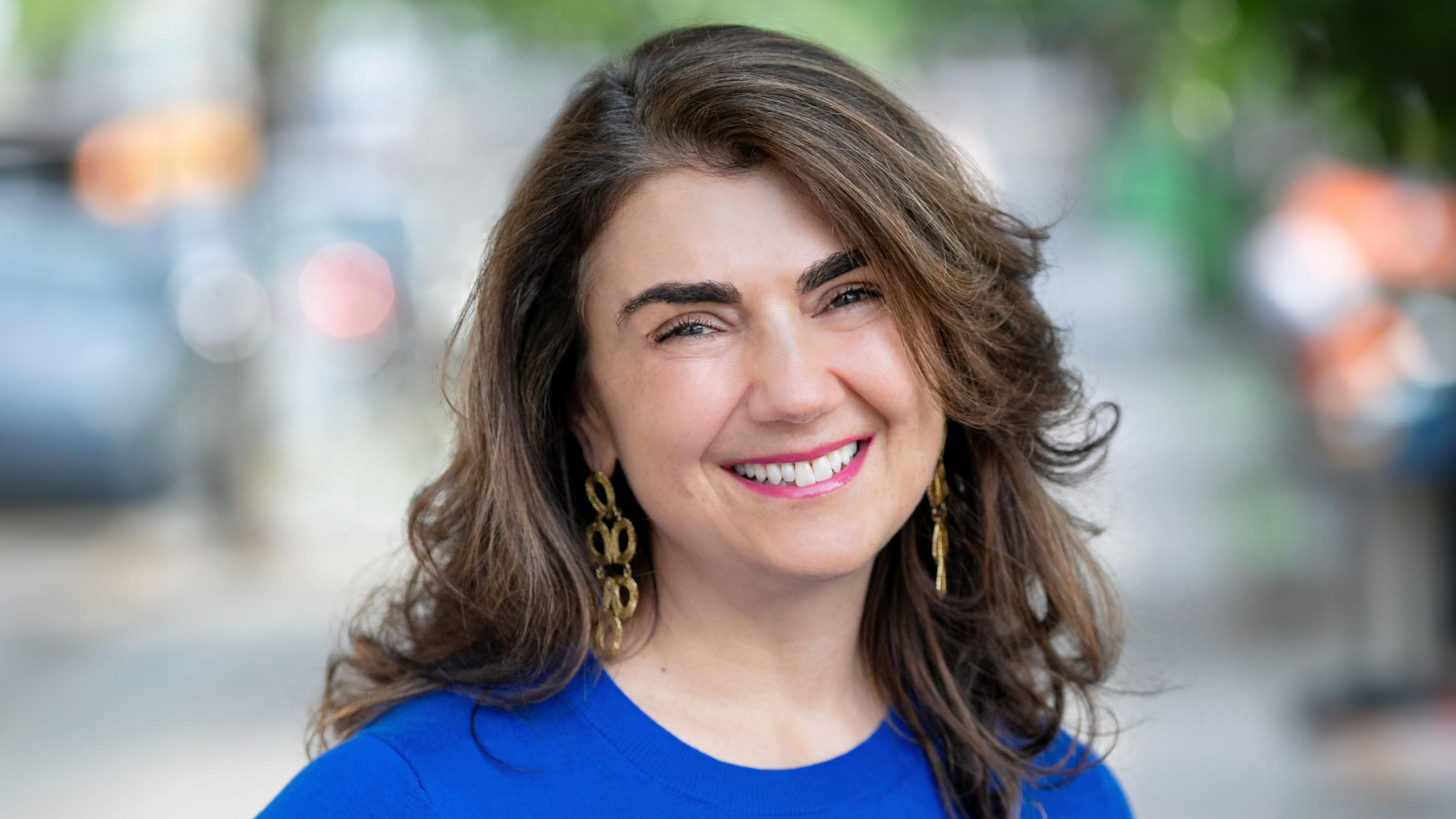 executive coach Alisa Cohn smiling, wearing a blue top, dangly earrings, wavy hair