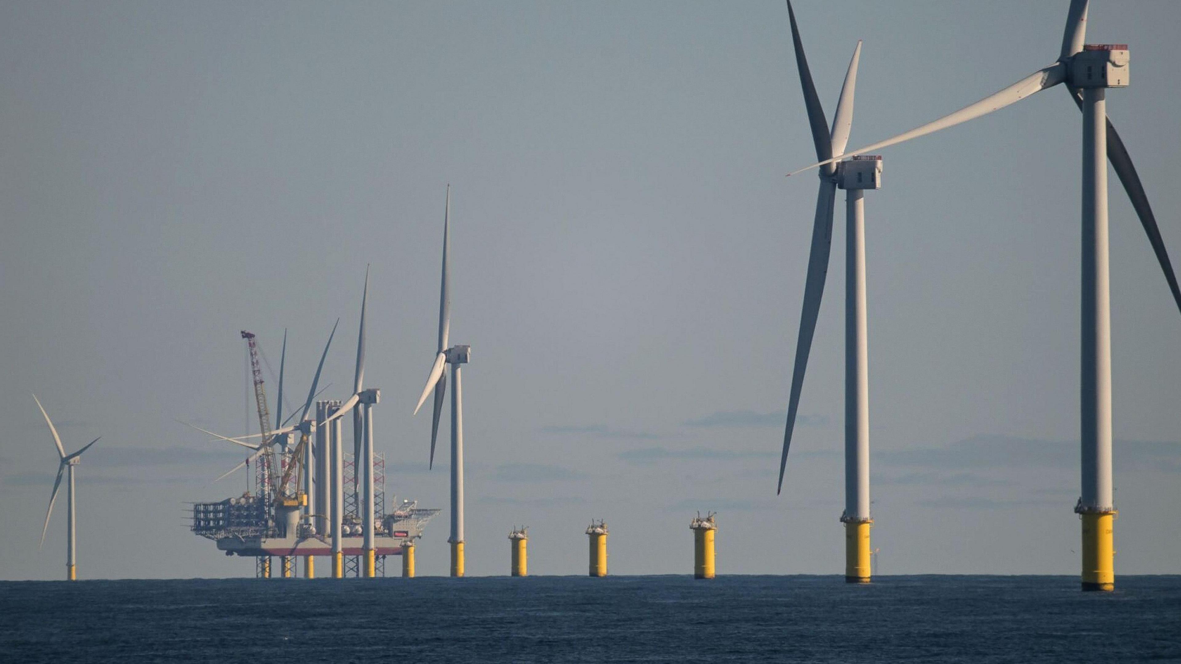 Photo of large offshore white wind turbines with the sky in the background. There are about six wind turbines in a row with a ship in the background also. The turbines are spinning. There are some clouds in the sky. 