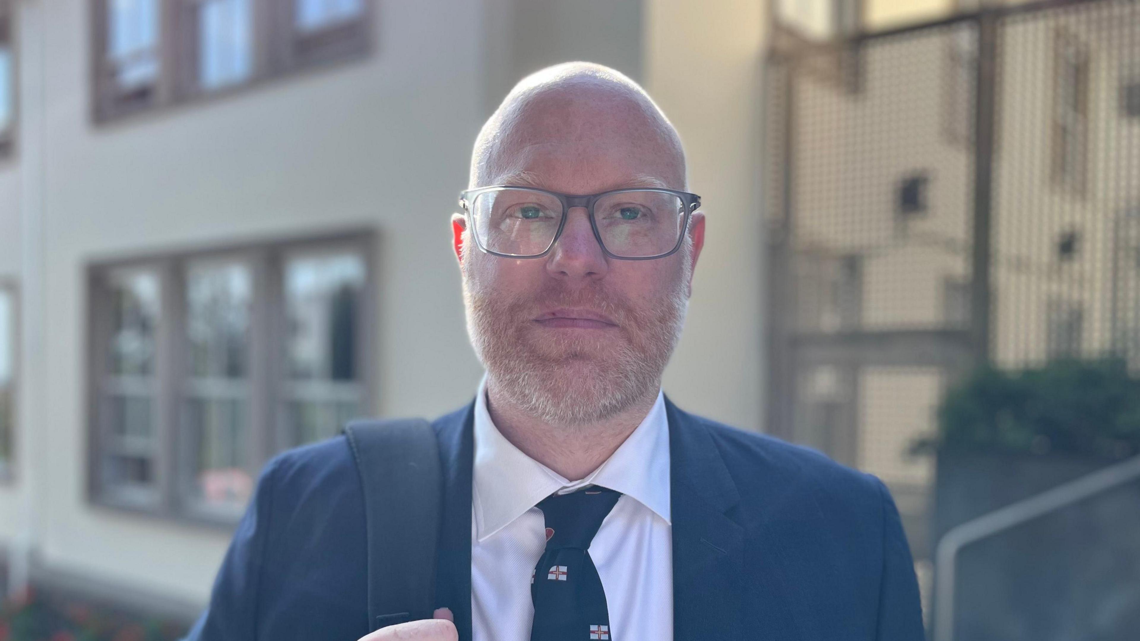 Deputy Rob Curgenven - A man with a bald head and ginger beard. He's wearing a navy blue suit and a white shirt, with a blue tie with Guernsey flags on it. He has spectacles on and a bag strap over his right shoulder. 