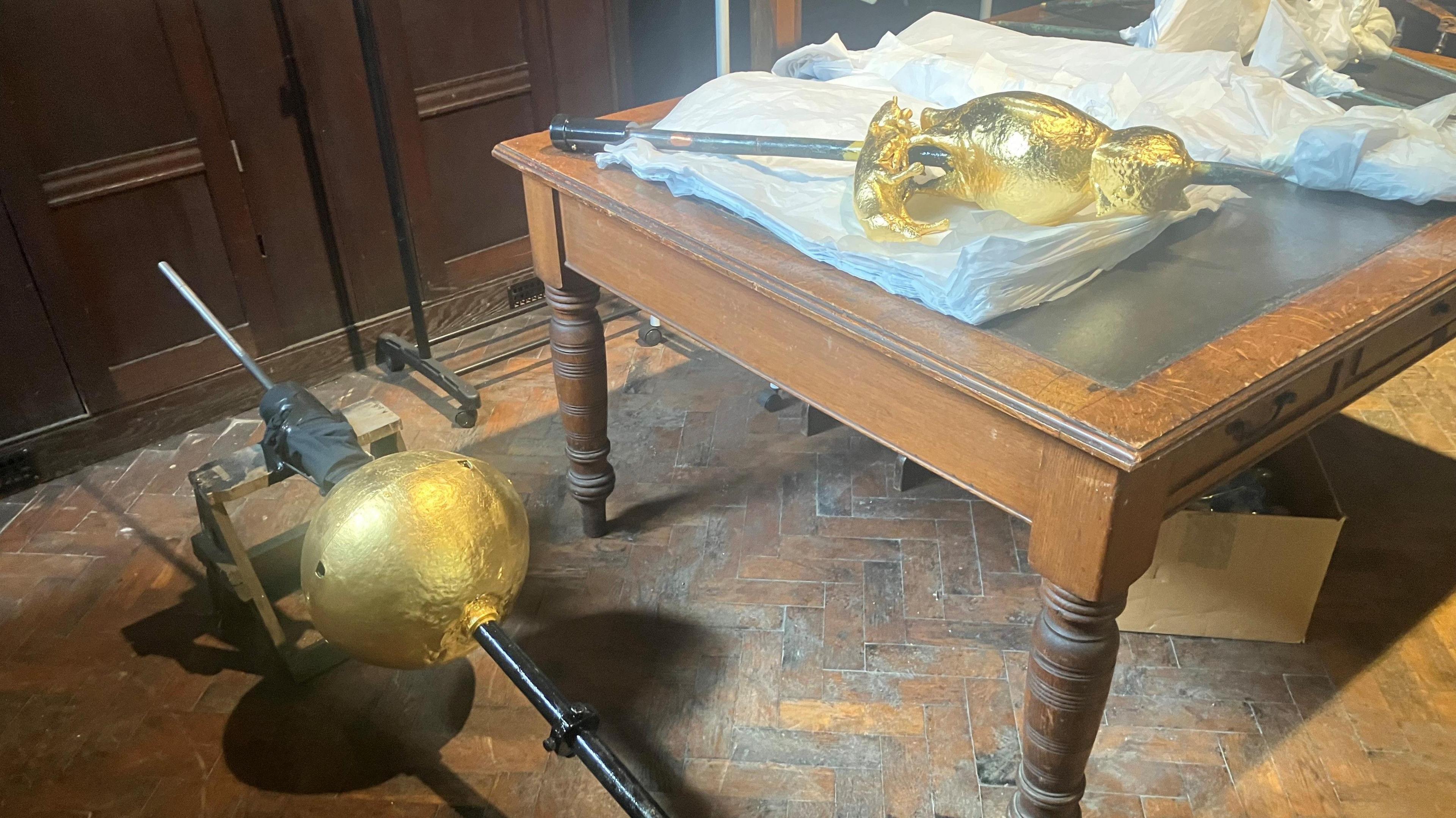 Photograph of the weather vane from St Wilfrid's Church in Standish. Part of it is lying on a table and another part is resting on the parquet floor.