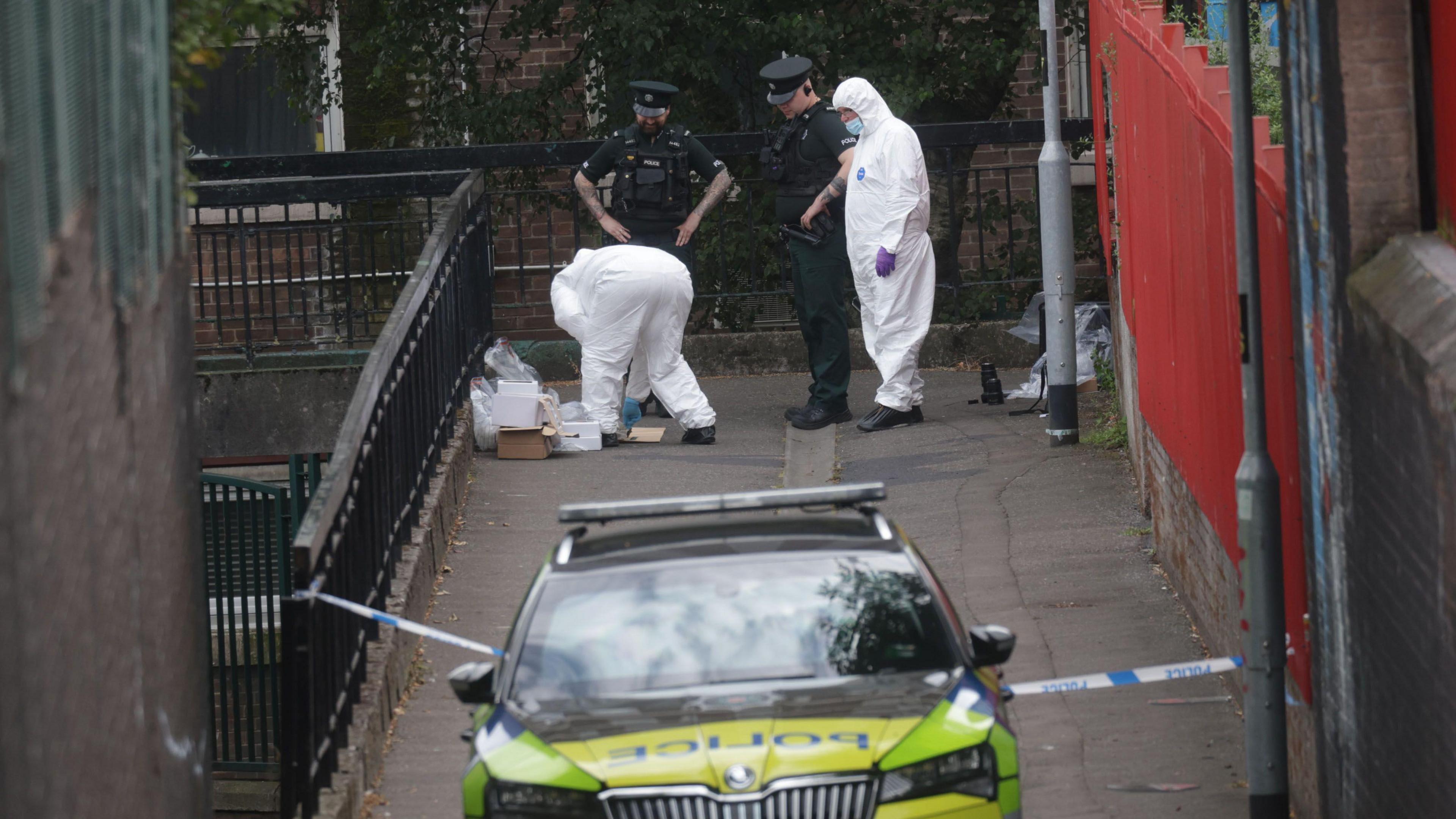 A PSNI car is parked in front of a walkway. Two PSNI officers are standing next to two people in forensic suits, one is crouched down sorting through carboard boxes and plastic bags.