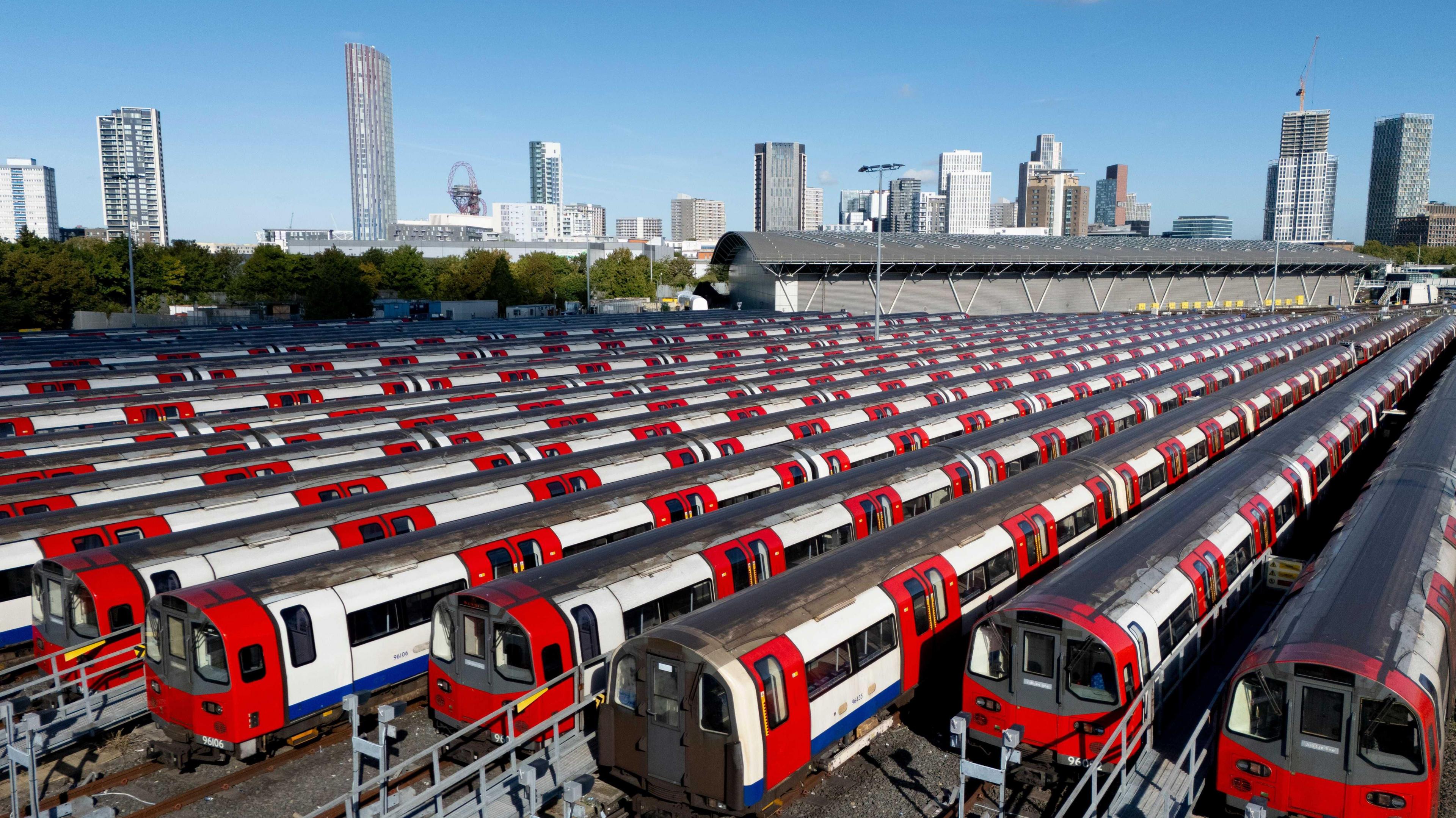 A row of Tube trains parked at a outdoor depot in London. Tall buildings are visible in the background.