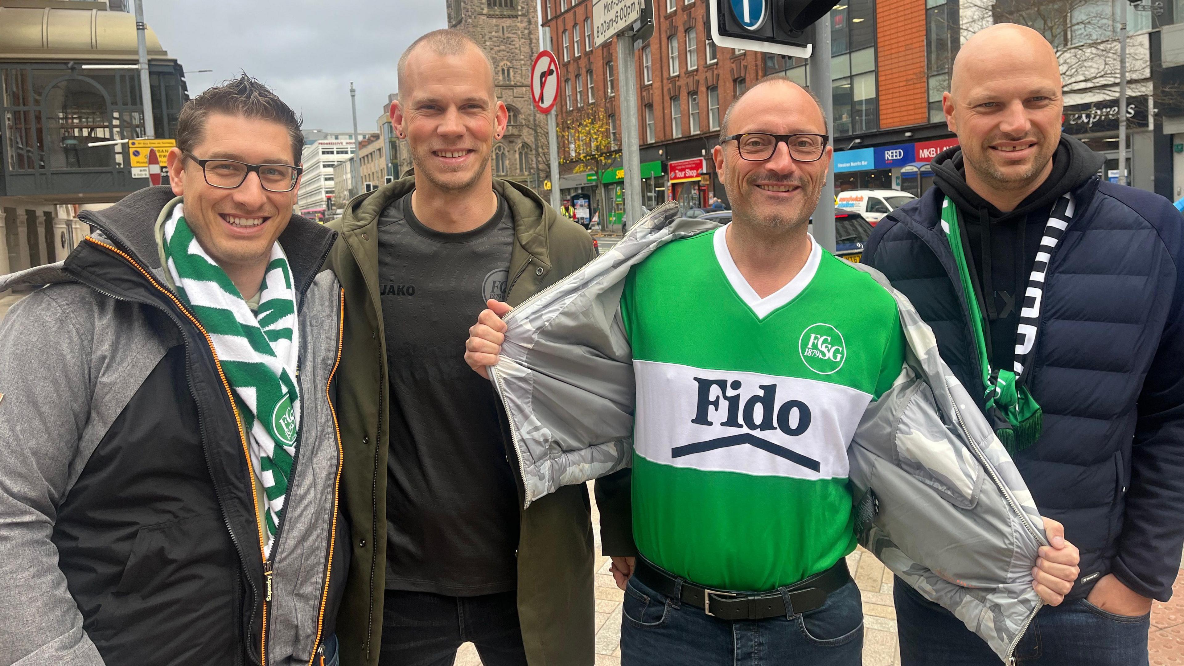 Four men stand on the street smiling at the camera. The man on the left wears a grey and black coat with a green and white scarf, the next wears a green coat and grey shirt, another man wearing square framed glasses holds open his grey coat displaying a green and white jersey. On the right, a man in a navy coat wears a black hoodie with a black and green scarf.