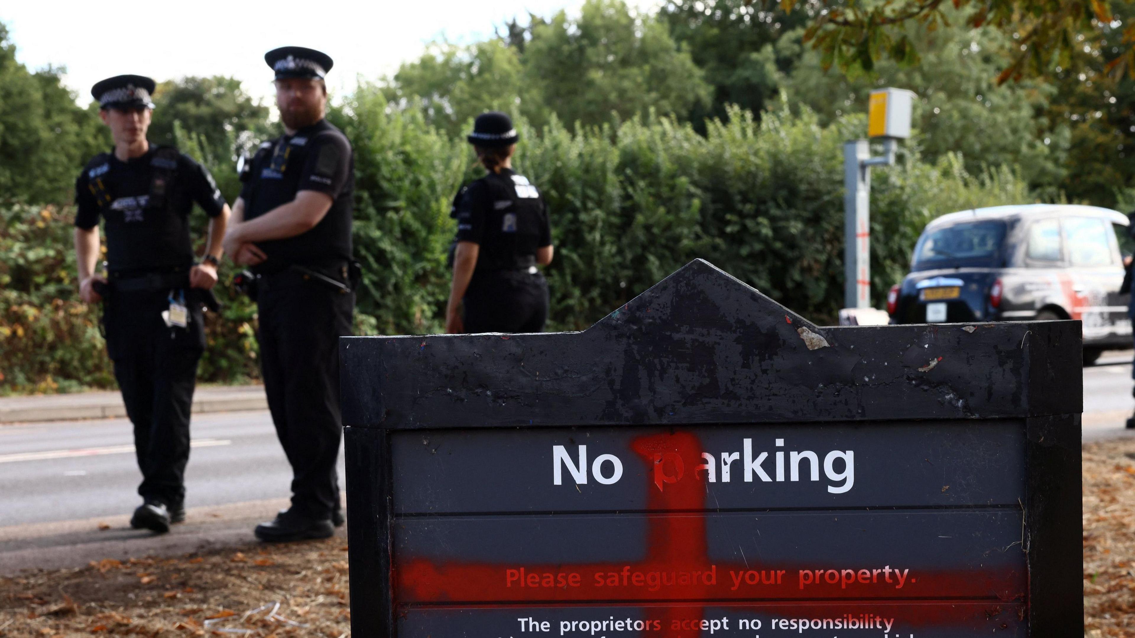 Three police officers in black uniform and hats stand on the pavement next to the hotel sign which reads "No parking". A red St George's cross has been spray painted onto it. Two of them look at the camera, an one has her back turned. There is a black taxi at the side and a speed camera.