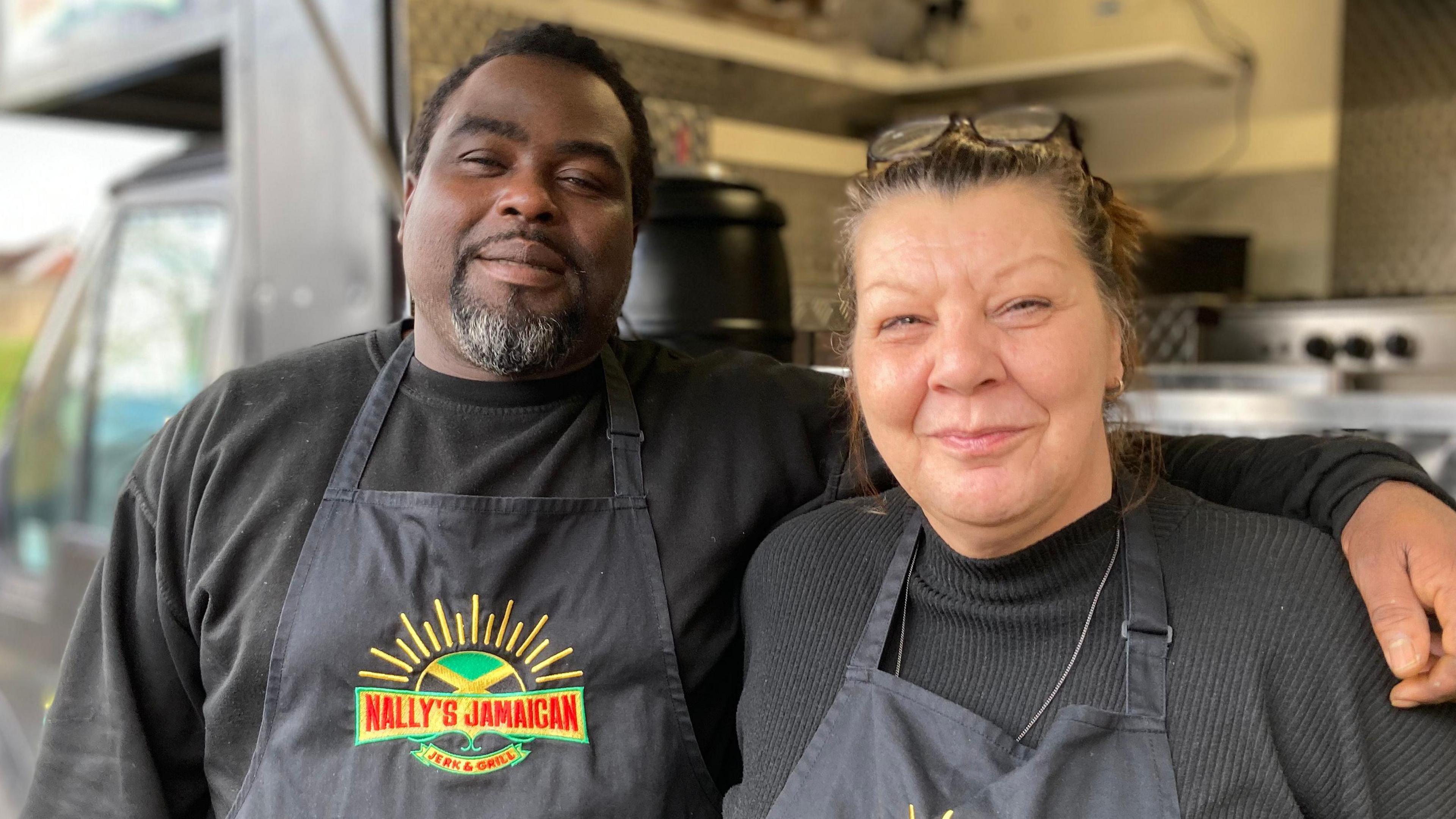 A man with with a grey goatee beard has his arm around a woman , they are both smiling toward the camera and are wearing black tops and aprons with red lettering on the apron which reads Nallys Jamaican Jerk & Grill. Behind them is a street food catering van.