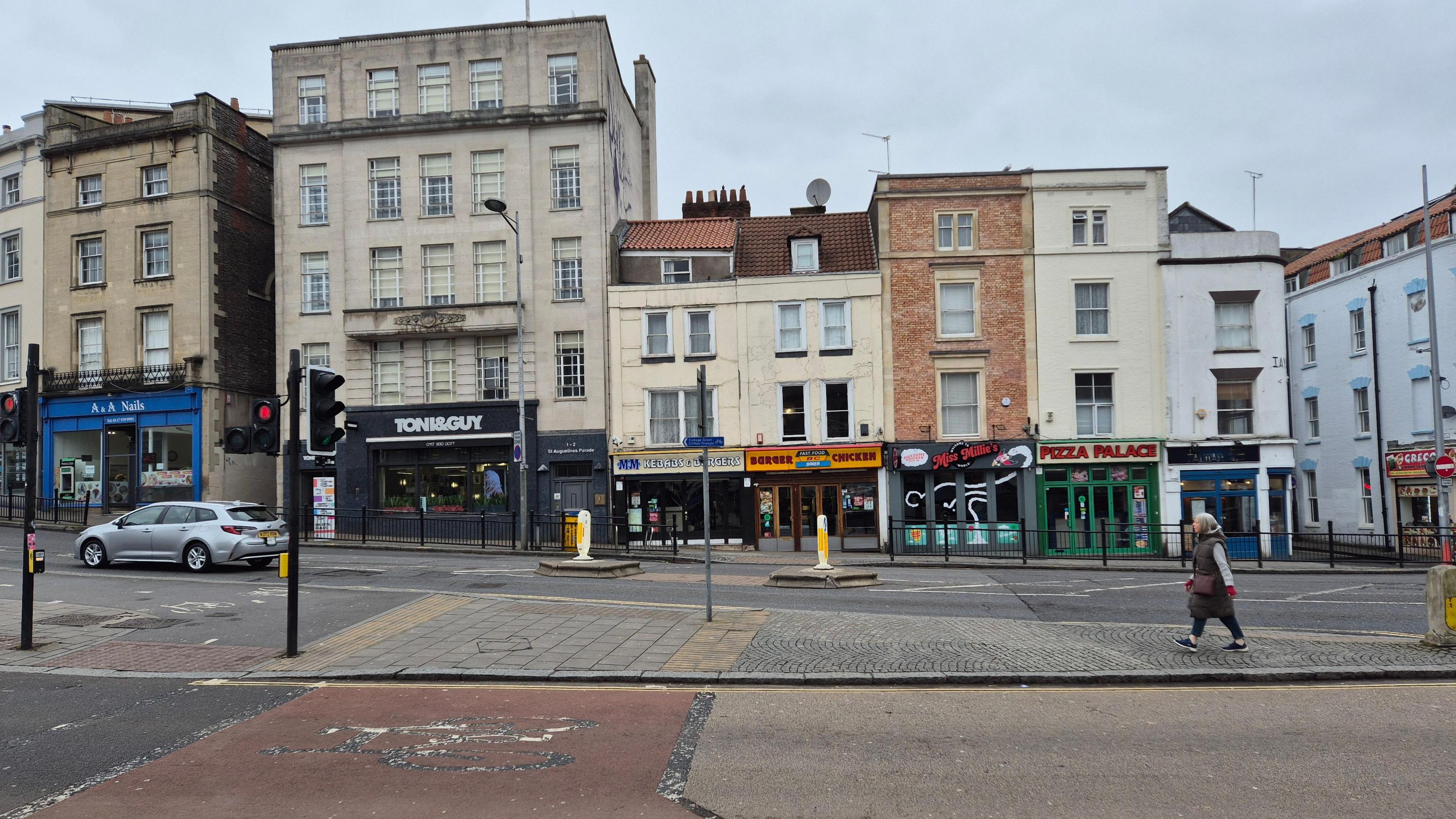 The pedestrian crossing on St Augustine's Parade near Anchor Road. There is a single pedestrian in the middle of the road and a car driving off. A row of takeaway shops is on the opposite side of the road - just up from Denmark Street.  