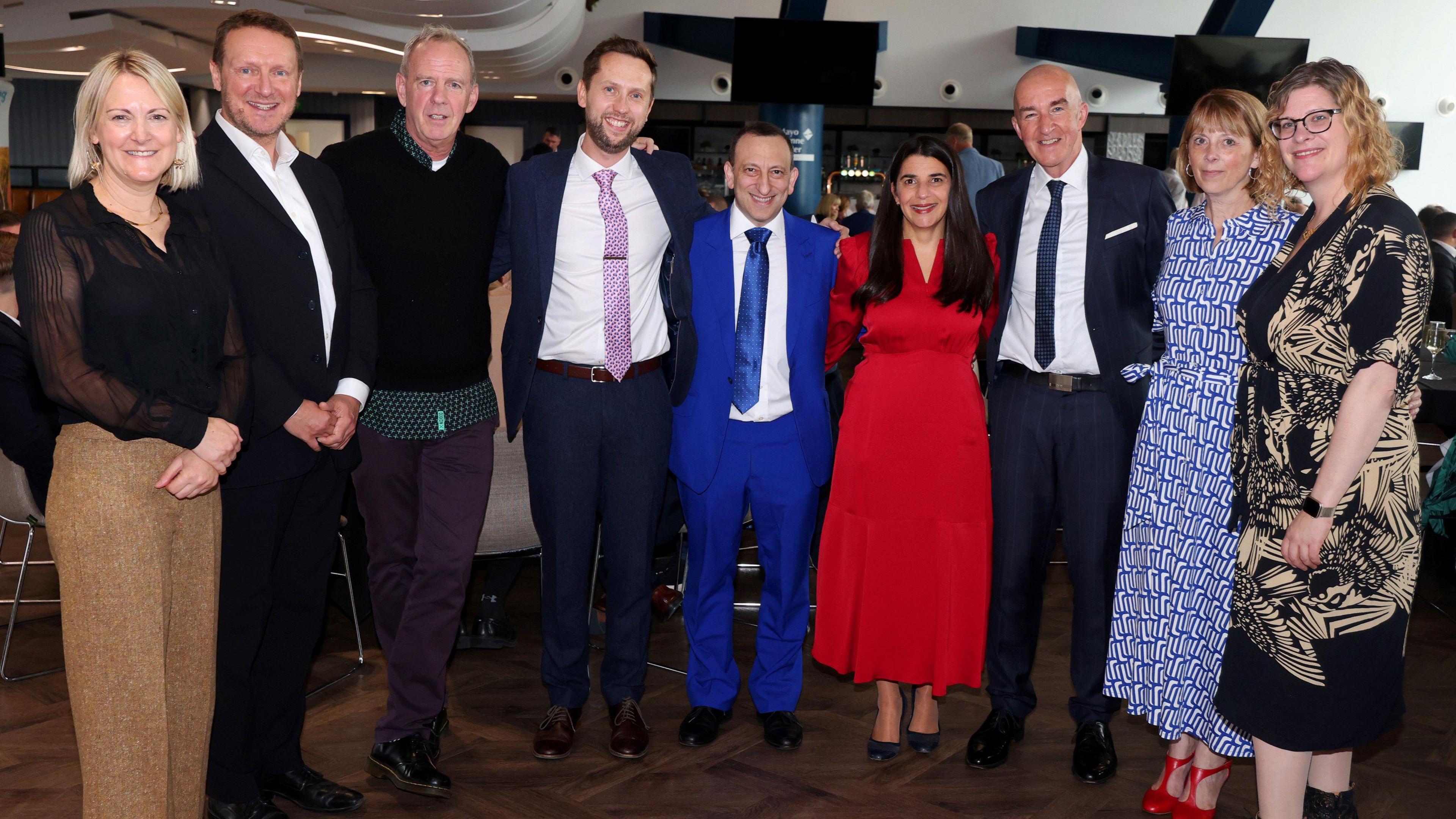 Nine people side-by-side looking at the camera. The group includes DJ Fatboy Slim in a black jumper and purple trousers, and Tony Bloom wearing a full blue suit with a blue tie.