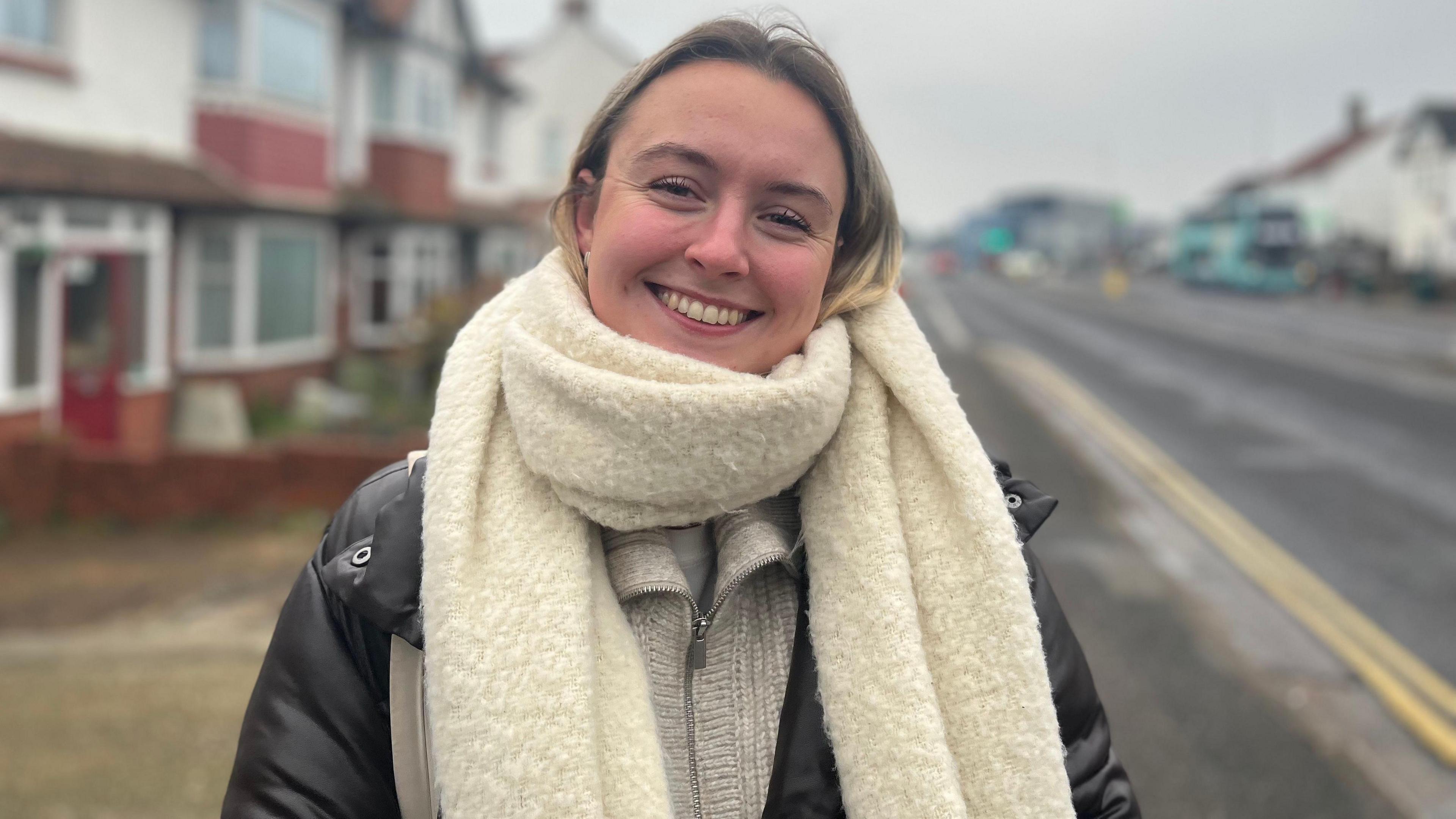 Elle De'Ath standing on a street wearing a large. fluffy cream scarf around her neck. She is smiling at the camera.