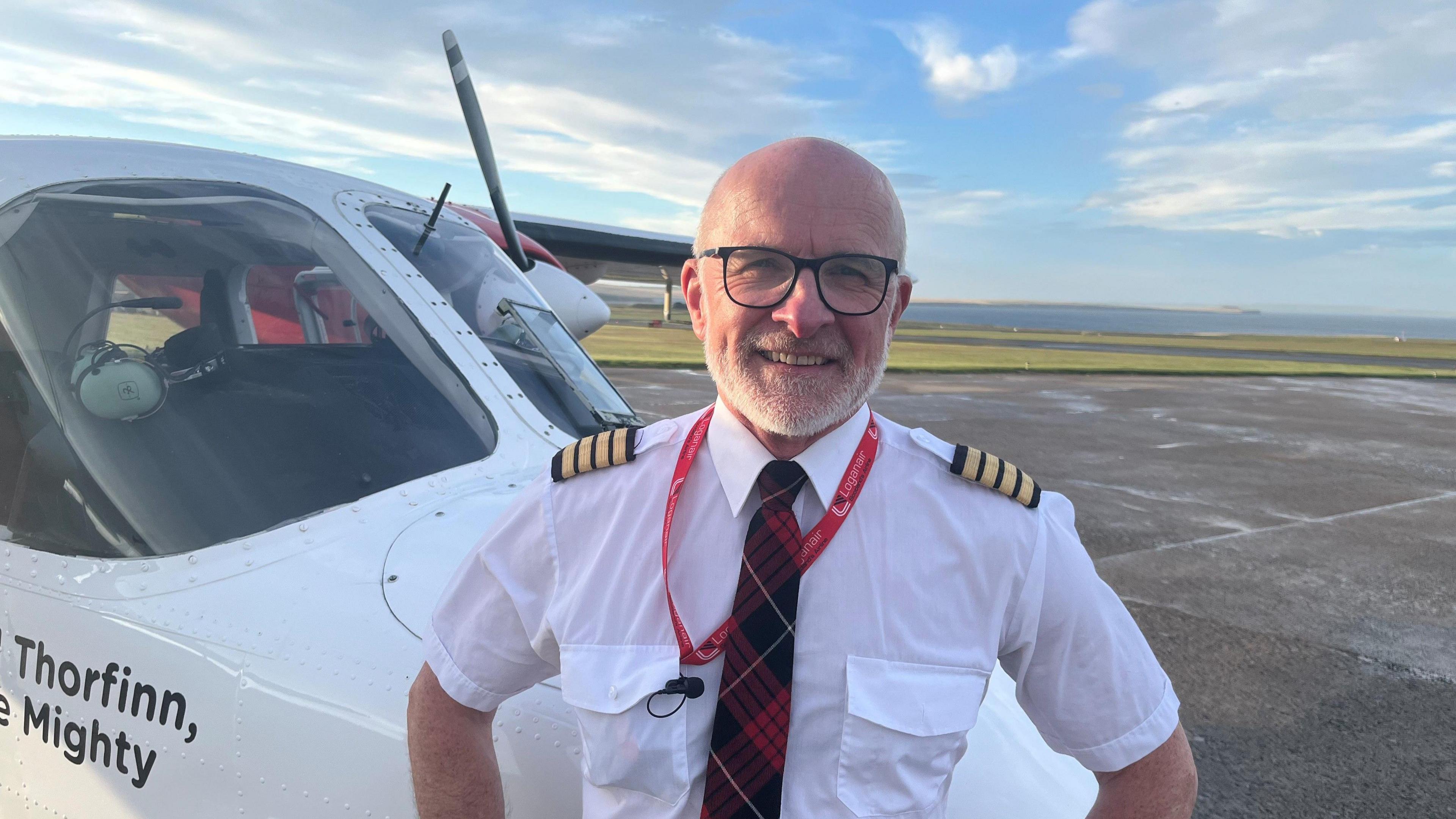 A male pilot, who is bald with a short white beard and is wearing a white shirt and a red tartan tie, stands next to a small aircraft. He has his hands on his hips and smiles. It is a bright sunny day and the coast is visible in the background, beyond the tarmac.