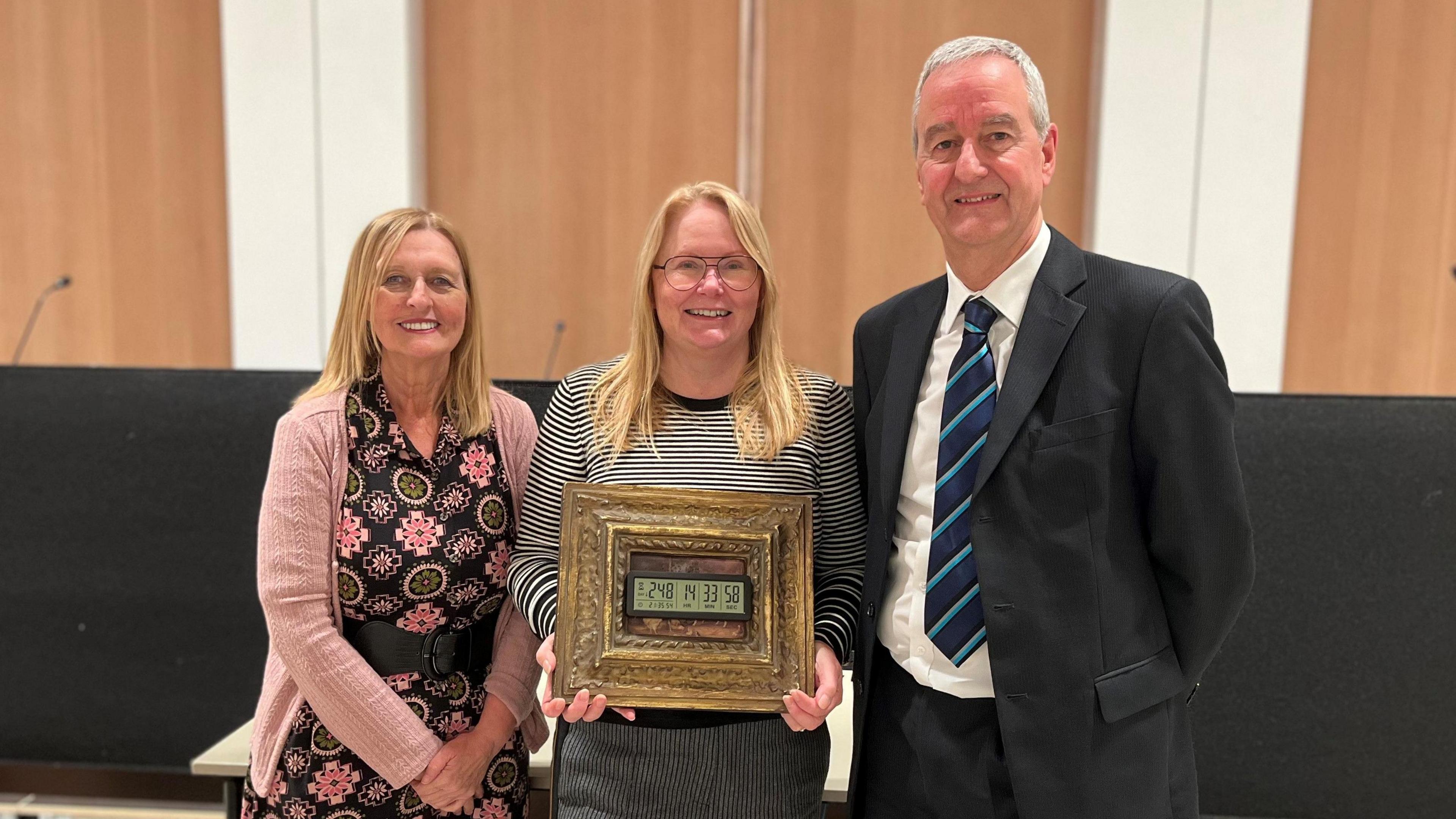 Campaigners Anne Loadman, Carolyn Basing and Nigel Taylor smiling at the camera in front of the council chamber's lectern. Anne and Carolyn both have straight blonde hair and blue eyes. Anne is wearing a black belted dress with green and pink flowers and a pink cardigan. Carolyn is wearing a black and white striped bloused as she holds a gold-frames electronic clock showing there were 248 days, 14 hours, 33 minutes and 58 seconds until the glass centre's closure. Nigel has short white hair and is wearing a black suit with a white shirt and a blue tie.
