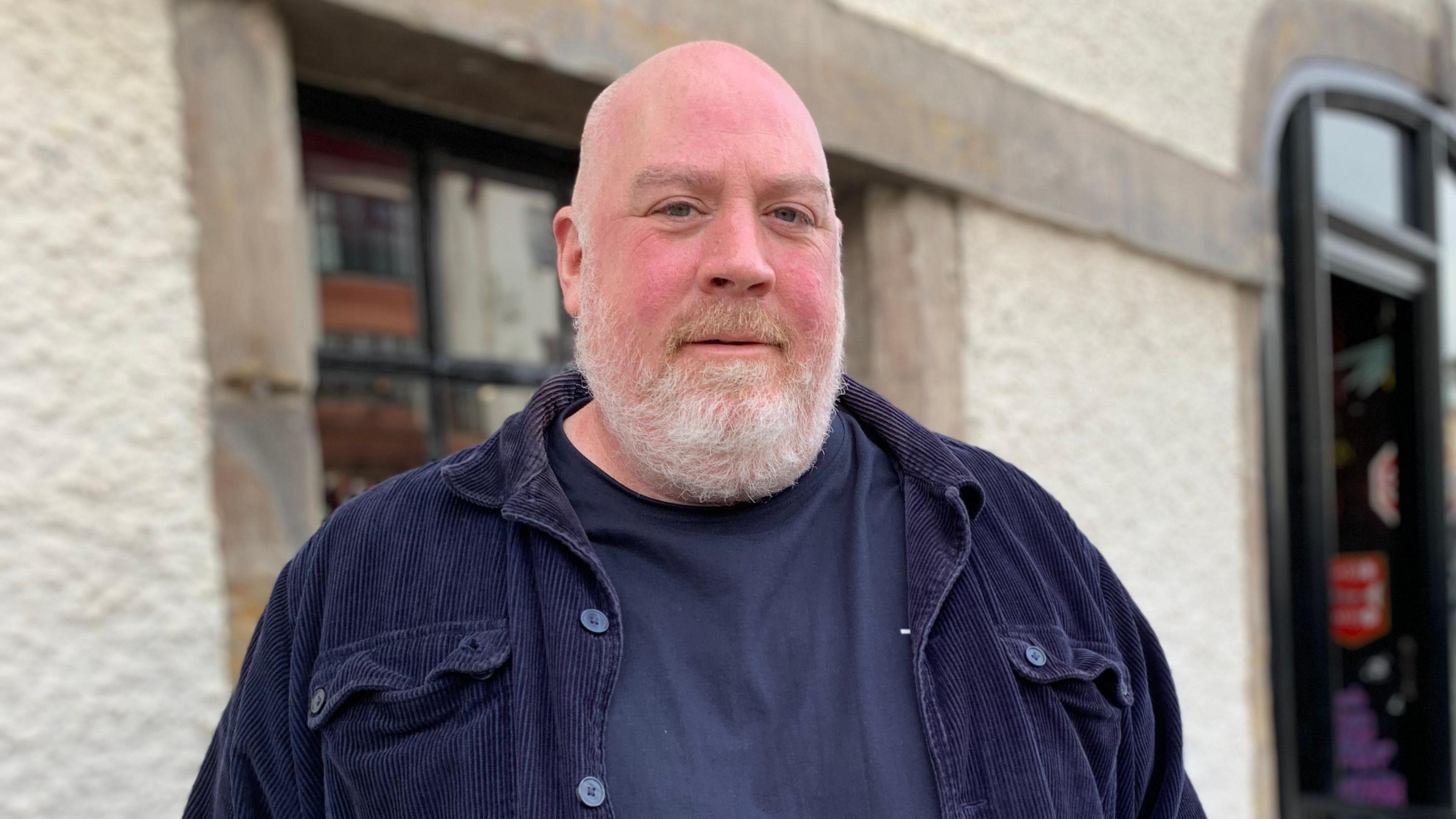 A bald-headed, bearded man in a t-shirt and dark jacket stands in front of a restaurant building