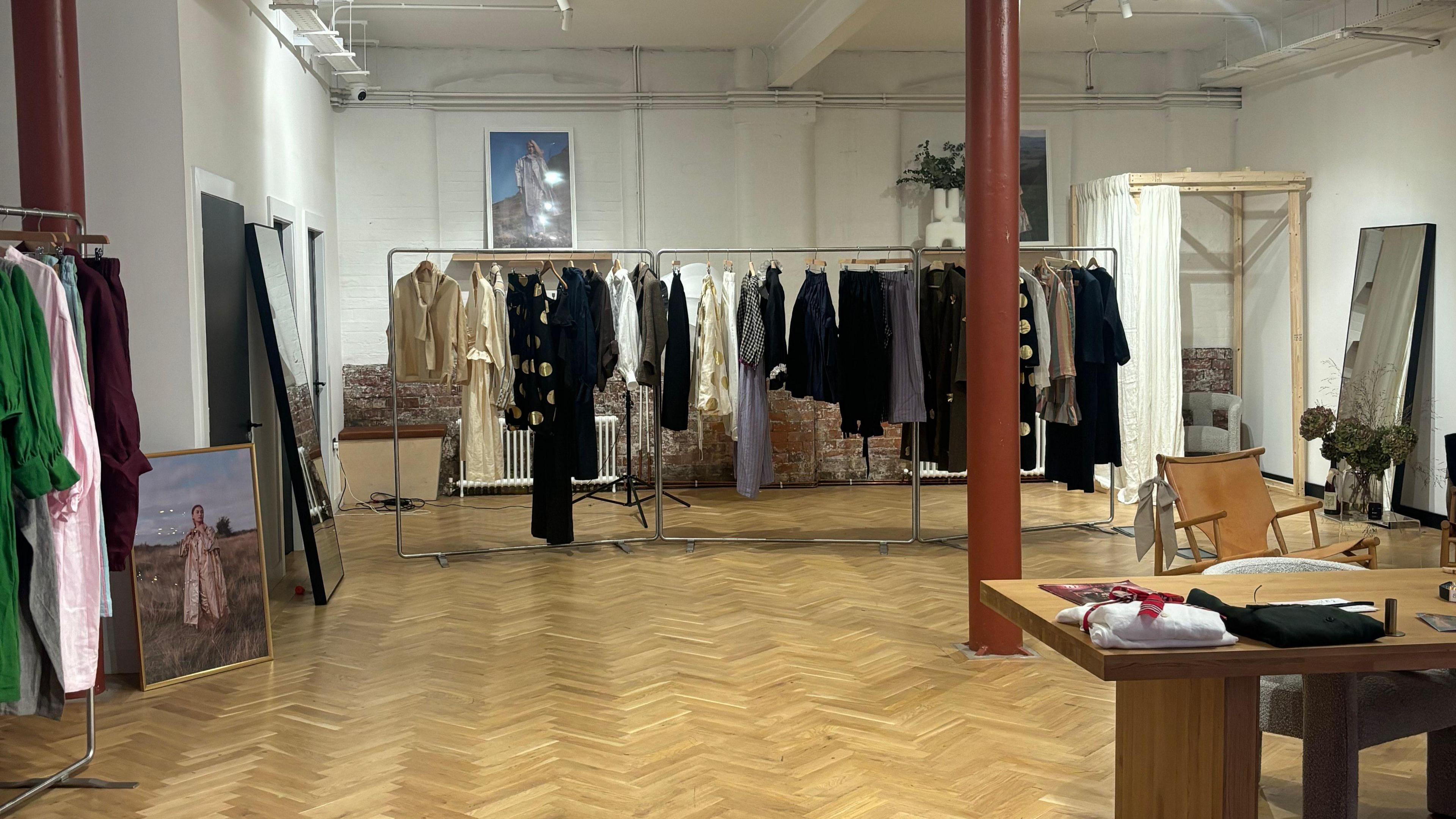 A view of the shop floor. Clothes are hanging on rails. There is a changing room in the corner. Two mirrors are on opposite sides of the floor. At the forefront there is a wooden table with items on top. The room is large with white walls and a red coloured pole running from the roof to the floor. The floor is wooden. There is also a picture of a woman in a field visible. 