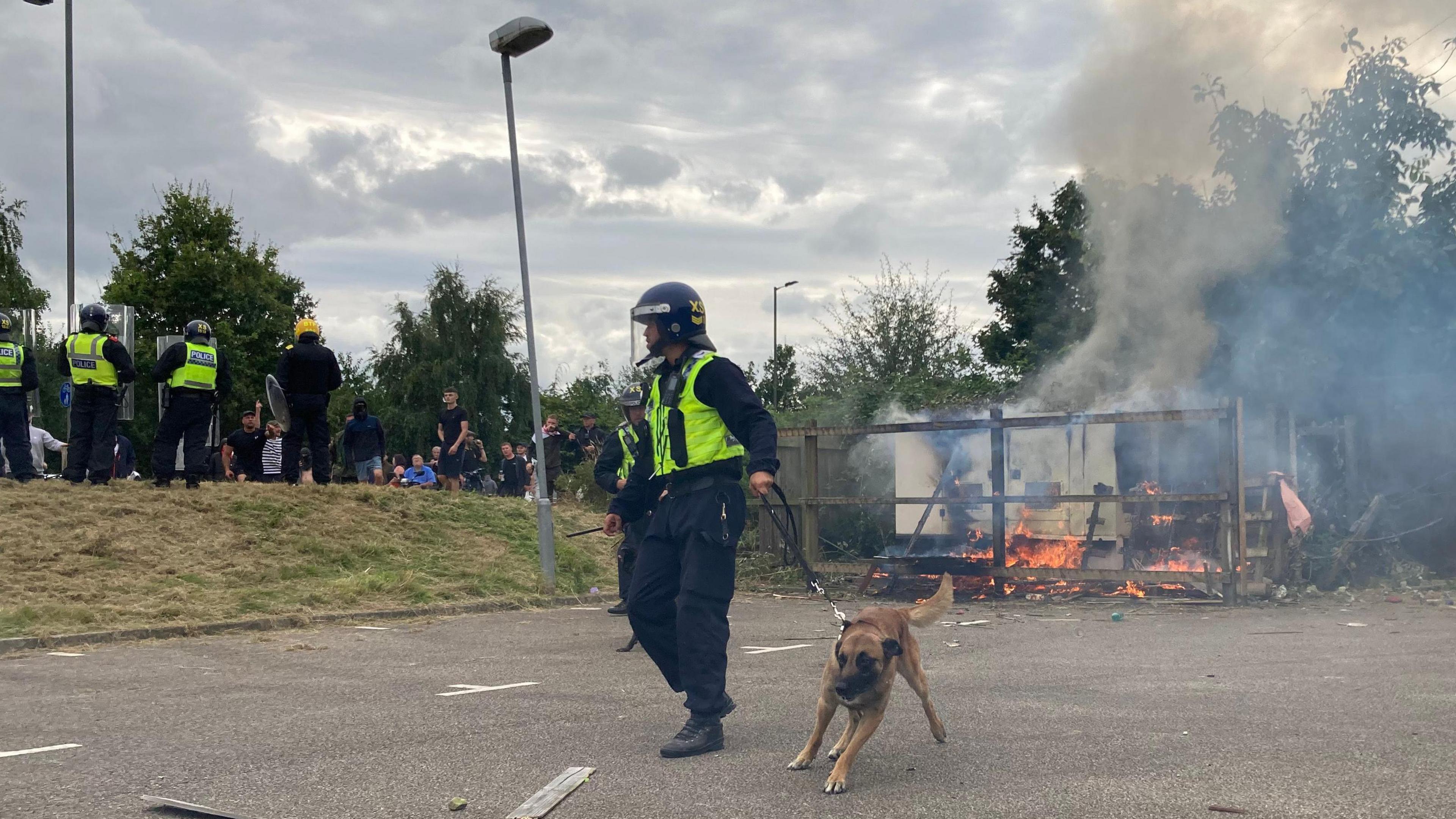 Riot police wearing helmets and holding shields at the scene of rioting in South Yorkshire, with one police officer in control of a police dog on a lead. A fire burns in the background, with smoke drifting from the scene. 