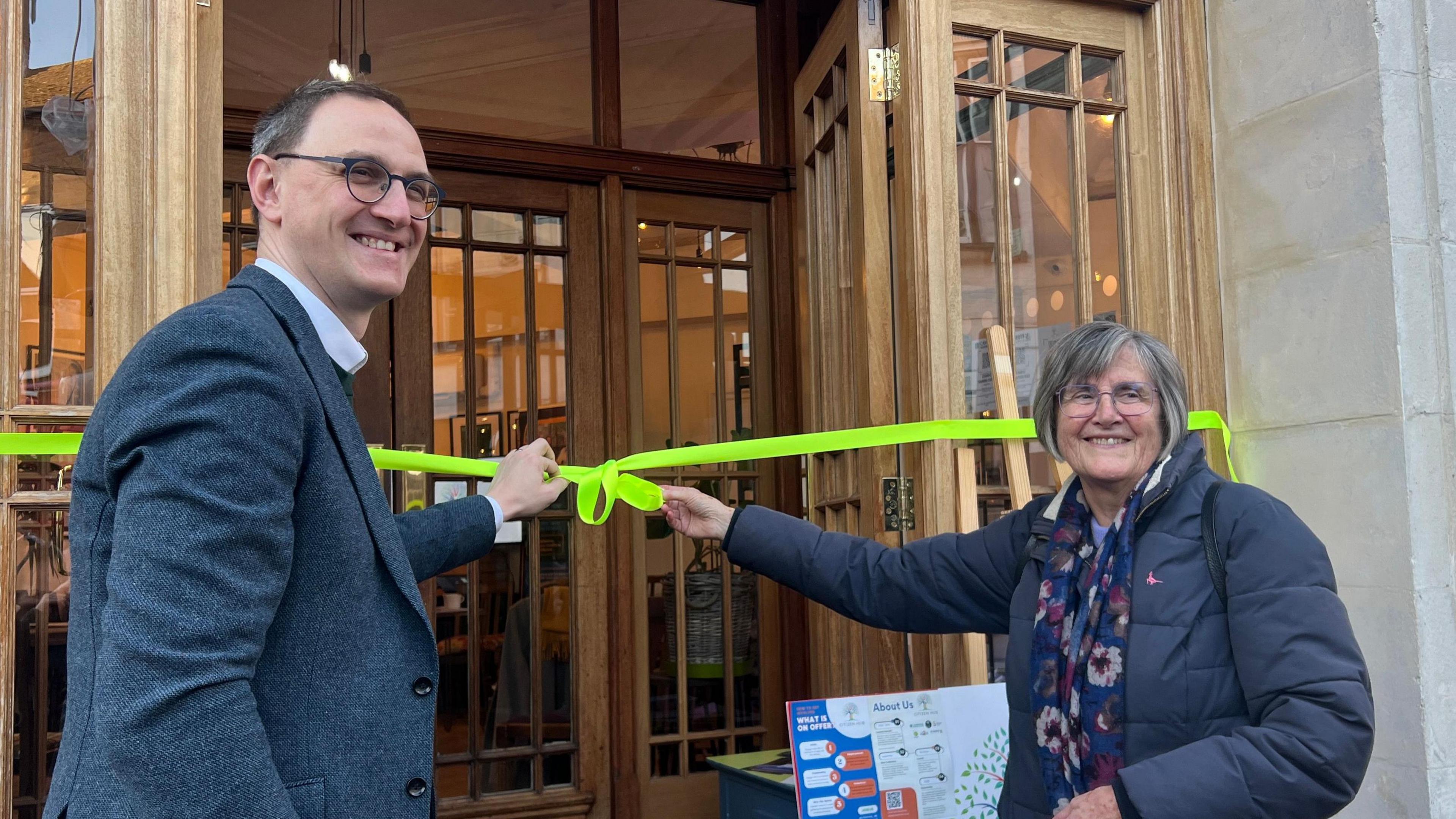 Ian Sollom and Mary Warren are standing in front of a bright yellow ribbon which stretches across the wooden doors of the renovated hub. 
Both have an arm reaching out to touch the ribbon and are turned at an angle to face away from the doors and to the camera. Sollom has round-frame glasses and and a blazer