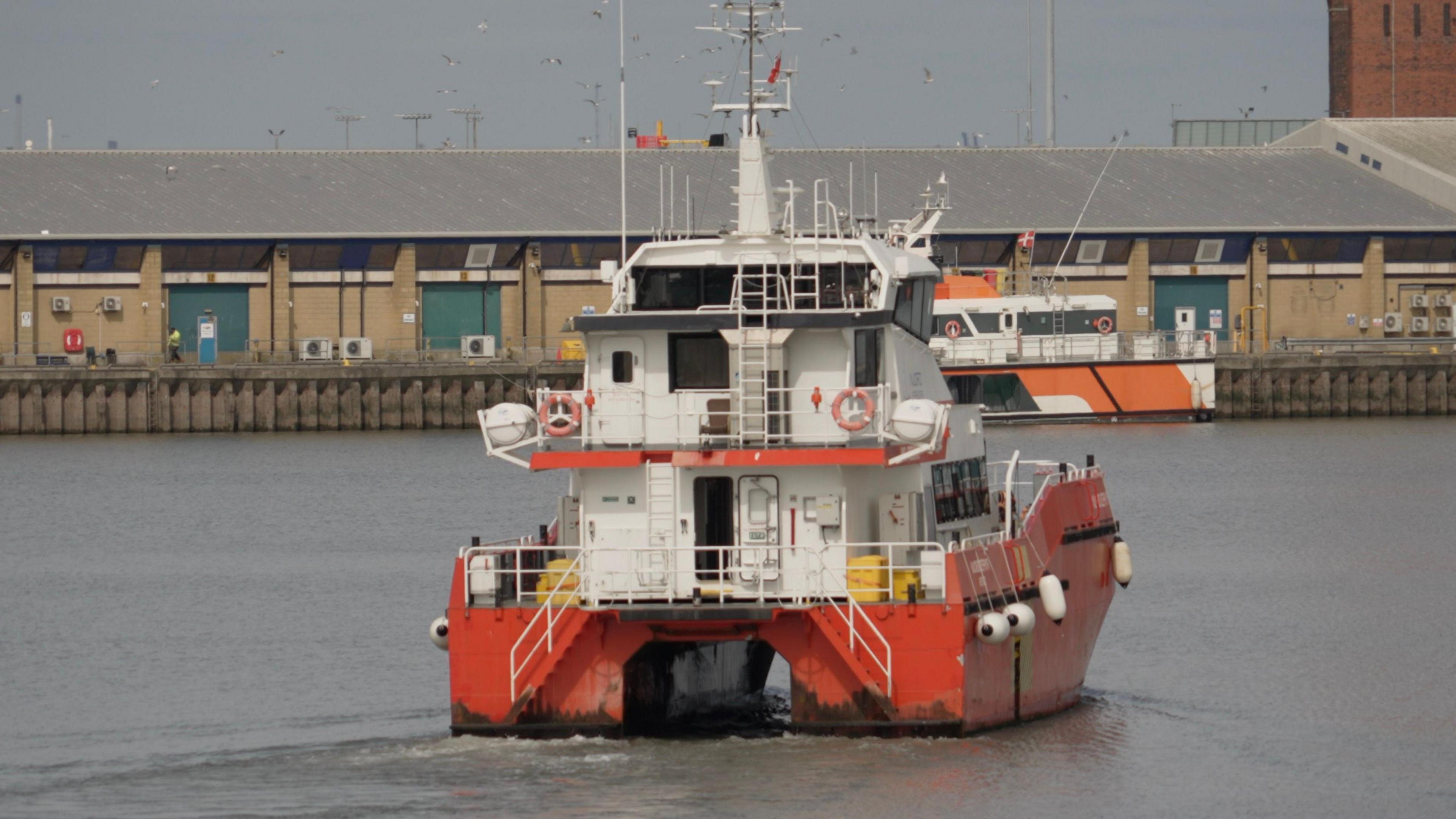 A white boat with a red base is sailing on water in a port, with buildings in the background