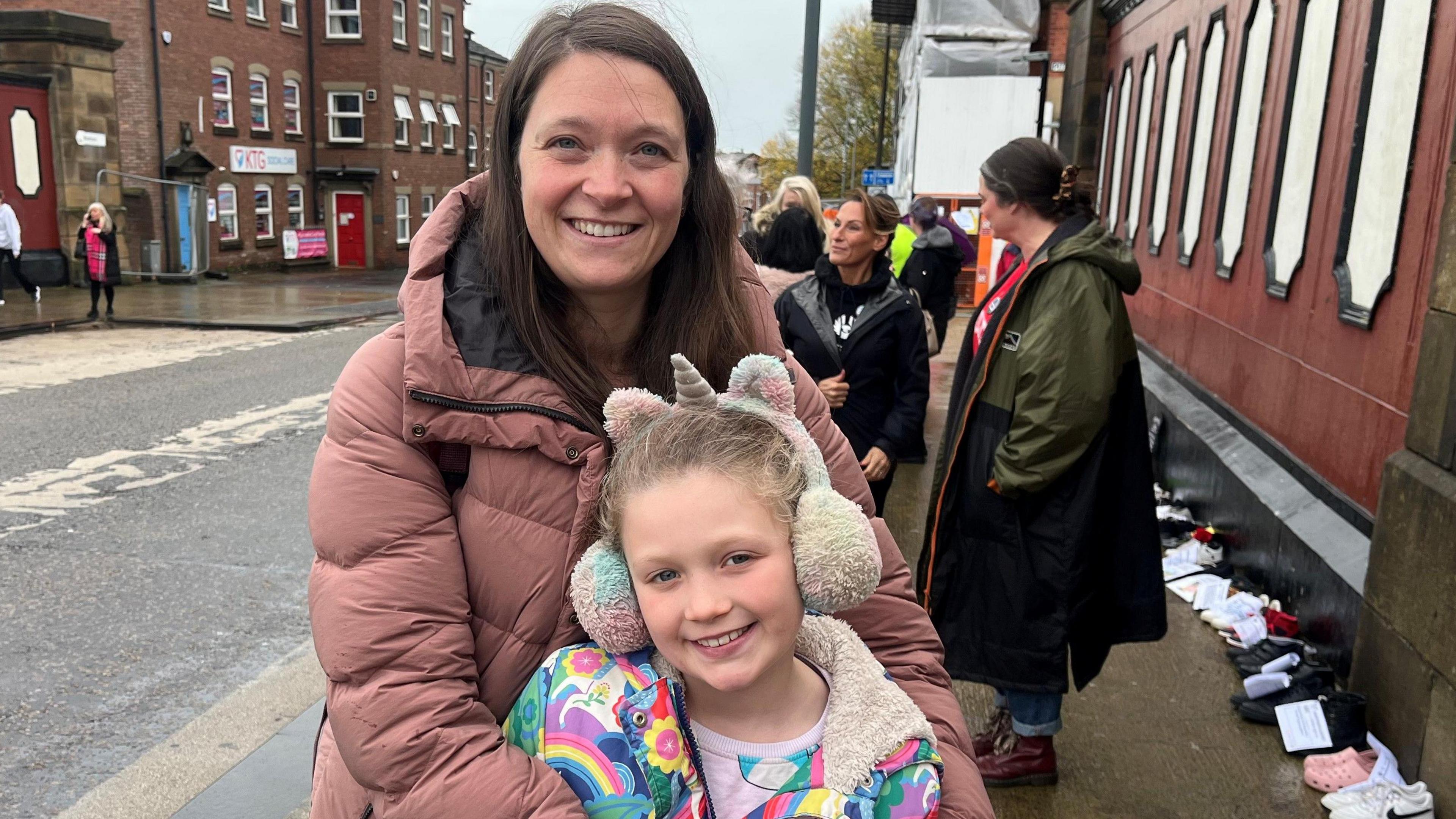 Sally Alker has a light brown coat, long brown hair and is standing in front of the building where the shoes are laid out.