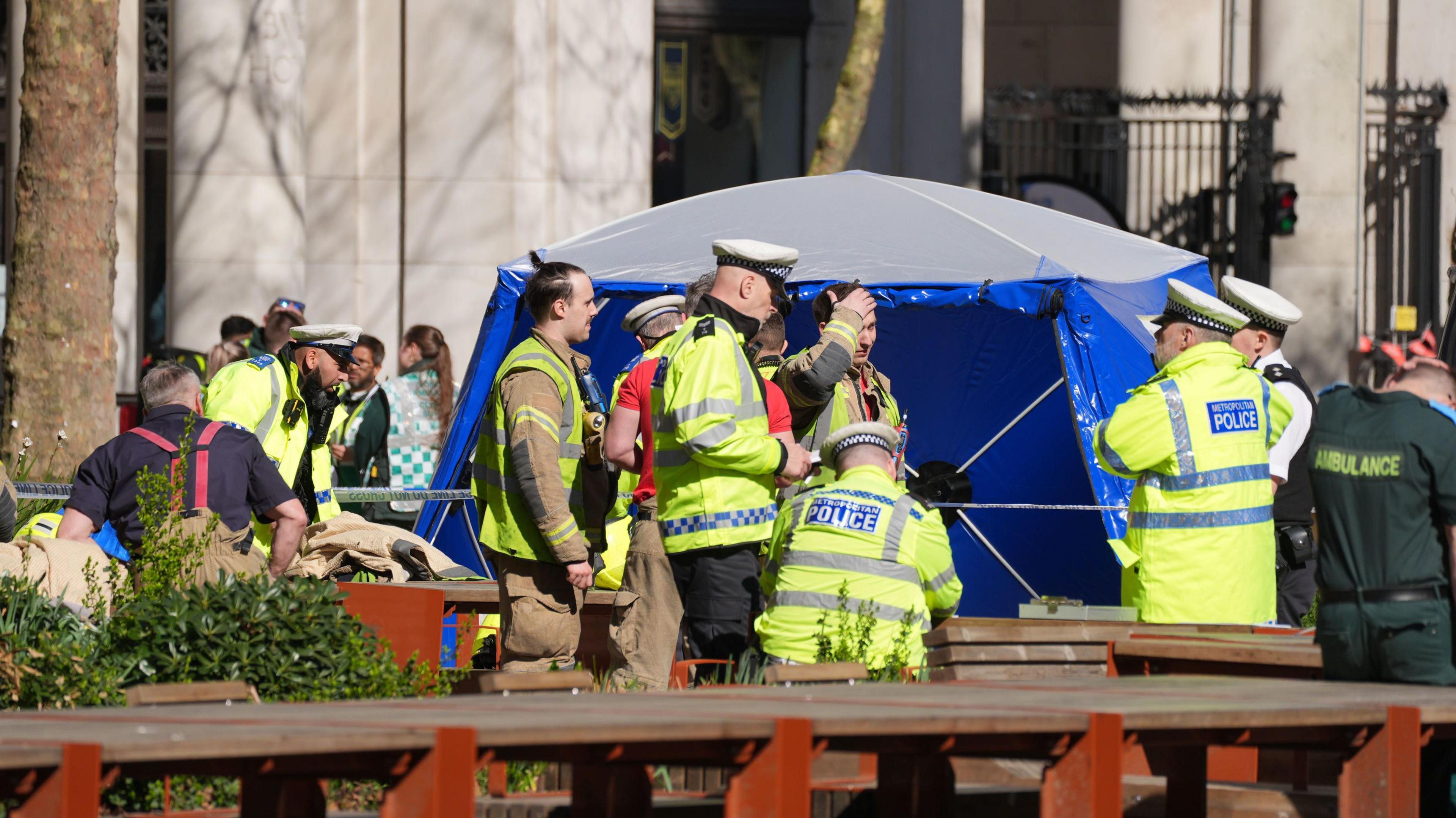 A blue and grey forensic tent is visible behind a group of 12 emergency workers including police, fire and ambulance staff, at the scene of the crash in The Strand