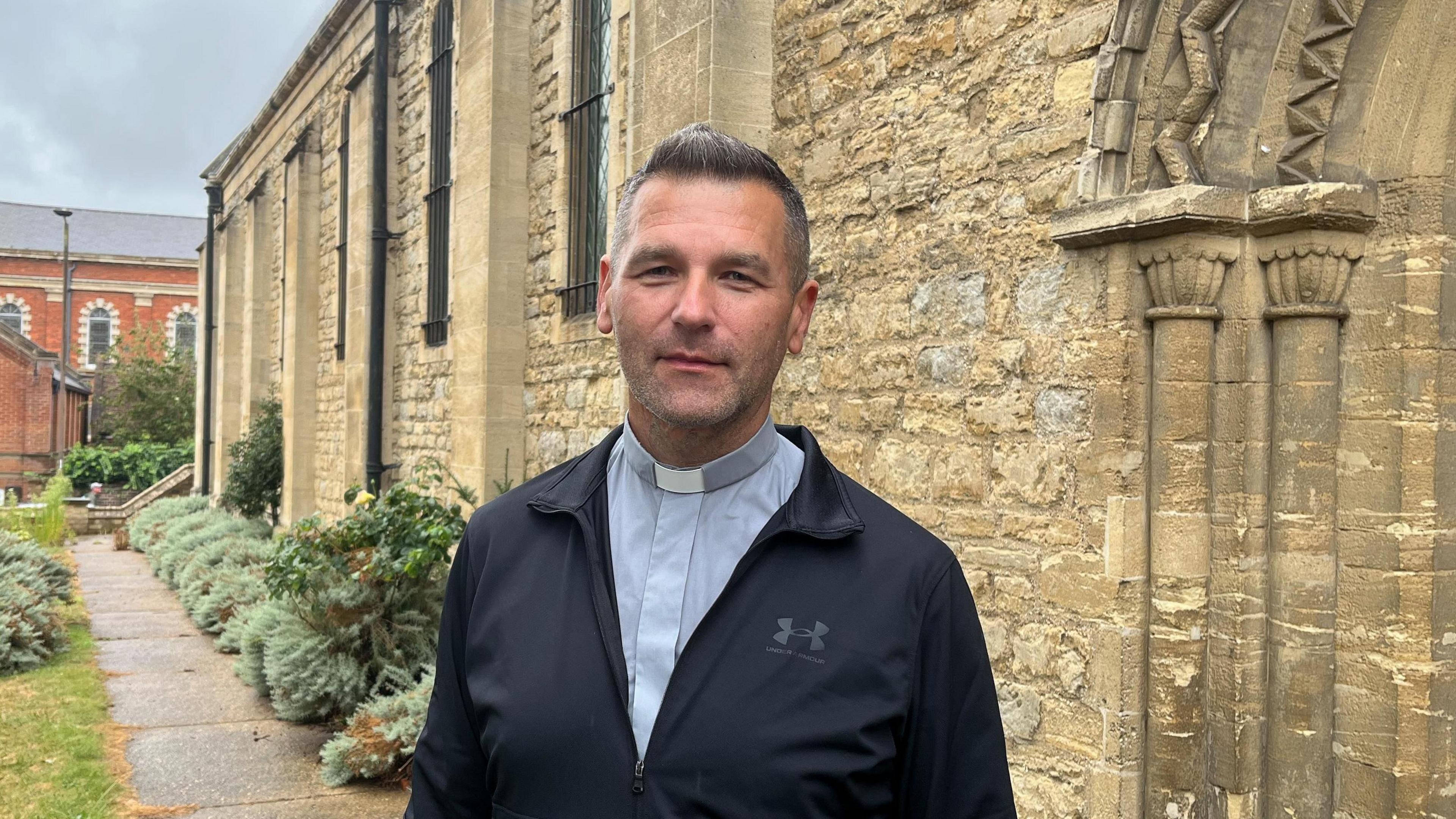A priest wearing a dog collar and a black jacket standing outside a church. He has light brown hair. In the background there are small bushes by the church.