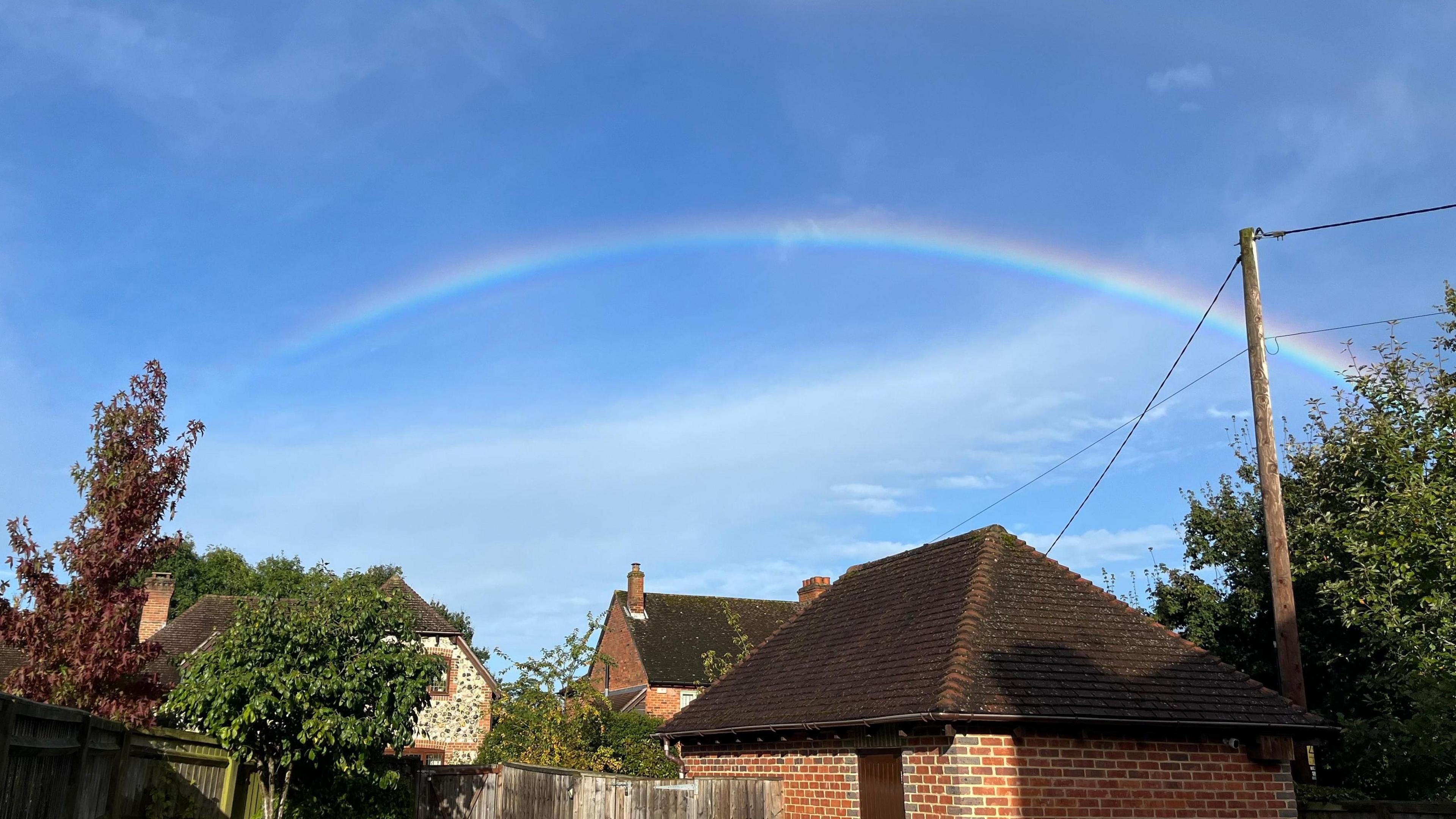 A rainbow shines in front of a bright clue sky over the rooftops of homes in Brimpton