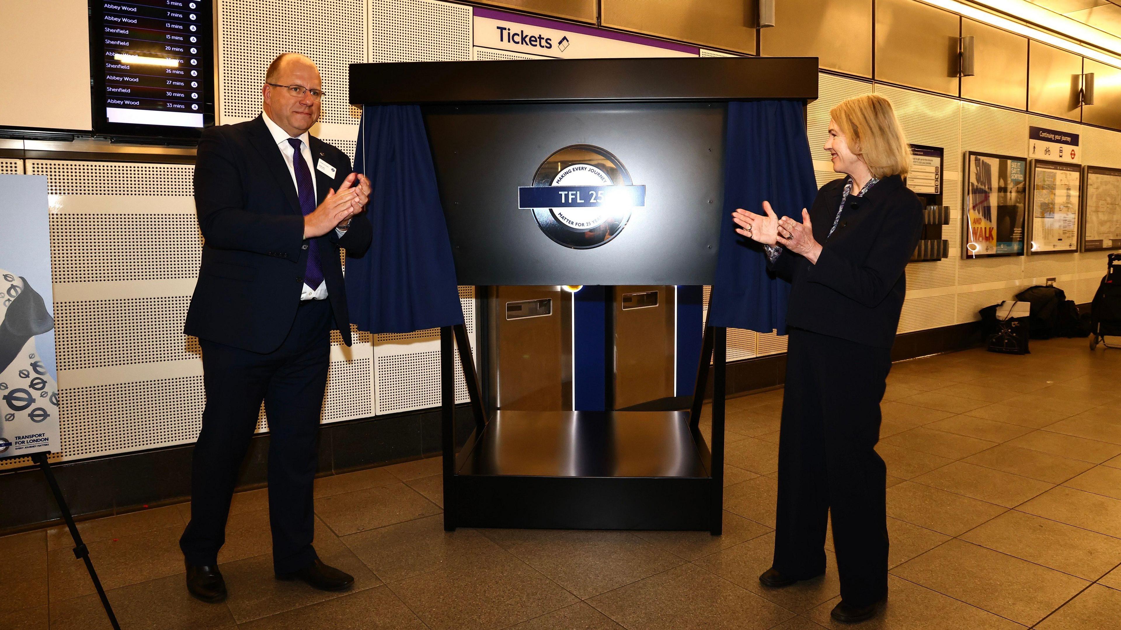 London's Transport Commissioner Andy Lord (left), a man with glasses wearing a black suit, white shirt and blue tie and London Transport Museum CEO Elizabeth McKay (right), a woman with short light hair wearing a black outfit, unveil a silver TfL 25 Roundel inside a TfL station